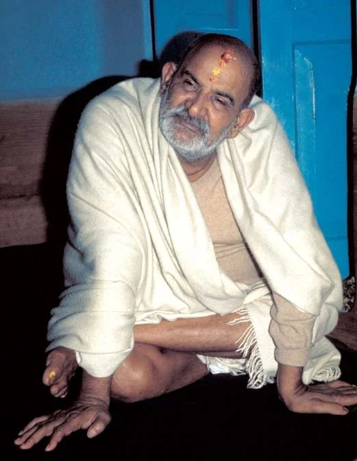 A man with a beard and mustache sitting on the ground with his legs crossed, wearing a cream-colored shawl and traditional clothing, with marks on his forehead and osculating the floor in a relaxed pose.