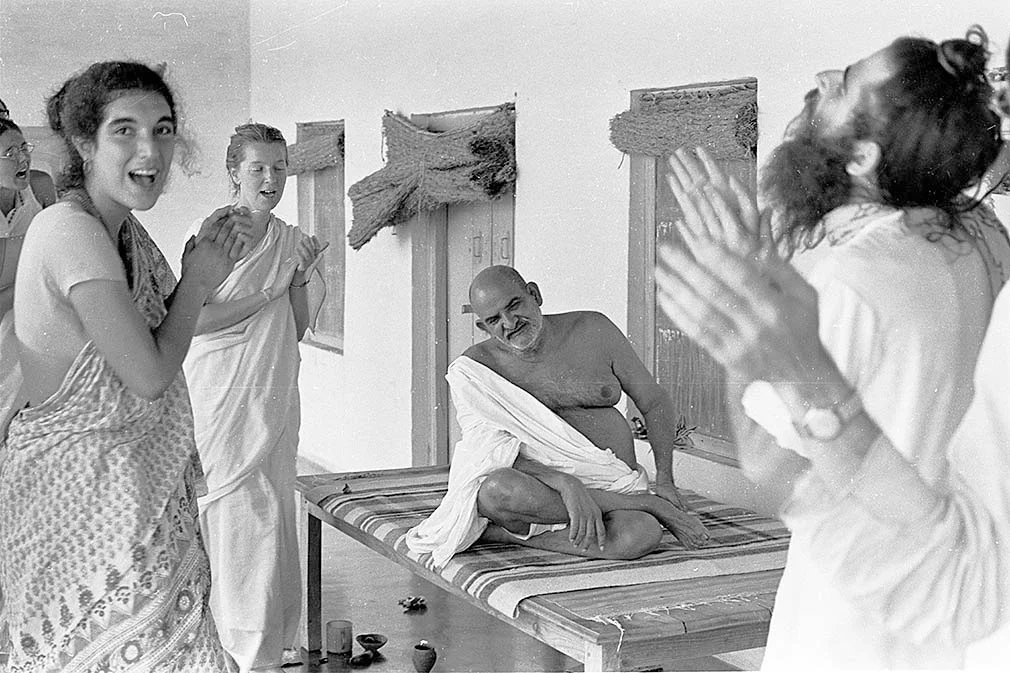 A group of women and a man are in a celebration, with the man seated on a bed and the women clapping and smiling, some with joyful expressions.