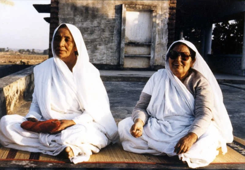 Two women wearing white traditional clothing and head coverings, sitting on a mat outdoors in front of a stone structure, smiling and relaxed.