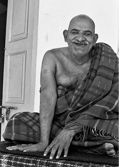 An elderly man with a mustache and bald head, sitting on the floor covered with a checkered blanket, smiling at the camera.