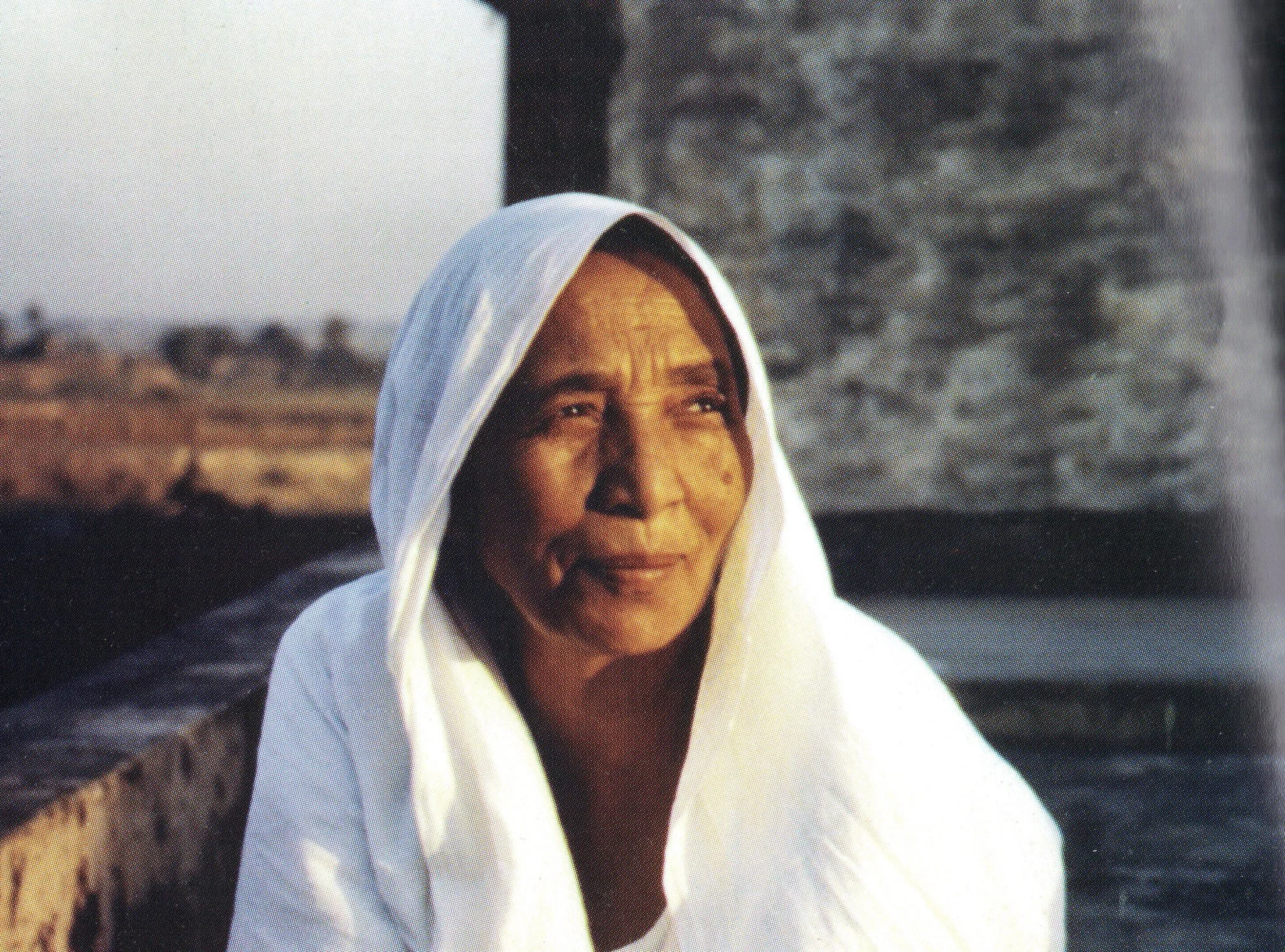 A woman wearing a white headscarf sits outdoors with a contemplative expression, with blurred landscape in the background.
