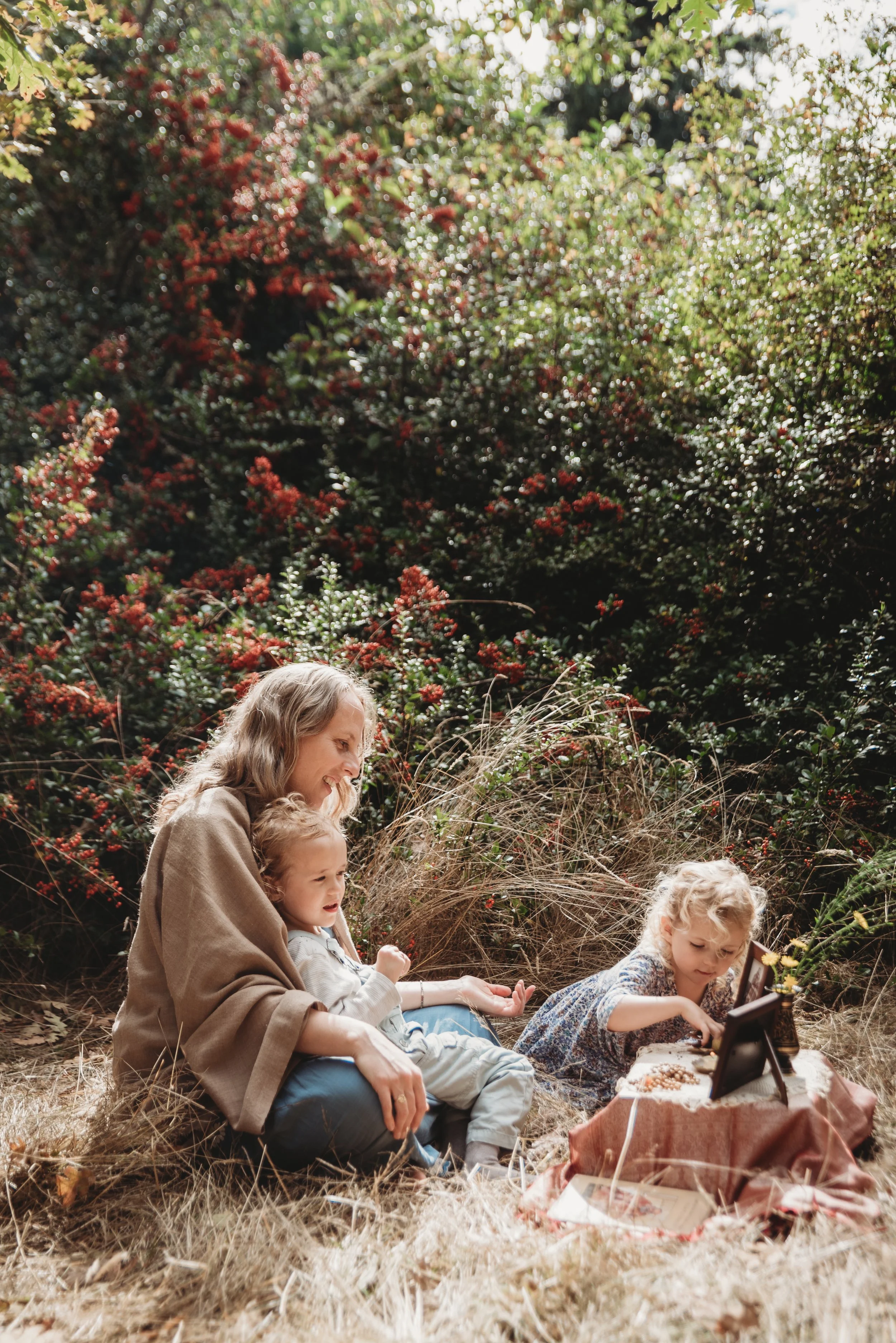 A woman and two young girls sitting outdoors on dry grass, with bushes and trees in the background. The girl on the right is playing a small piano, and the woman and girl on the left are watching and smiling.