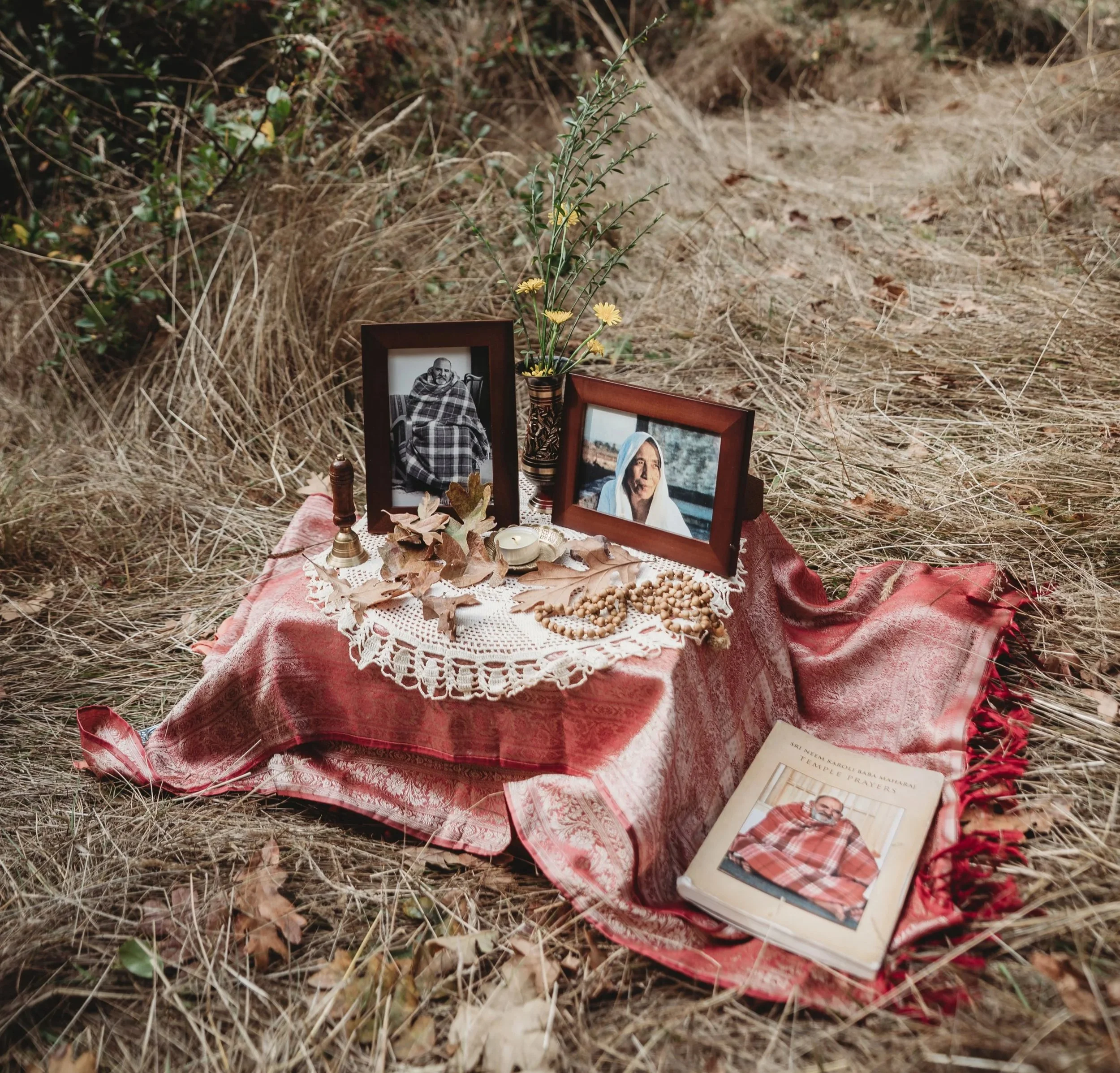 A memorial setup on a red patterned cloth with framed photographs of a woman and a man, a flower vase, dried leaves, a small bell, prayer beads, a candle, and a book, placed on dried grass.