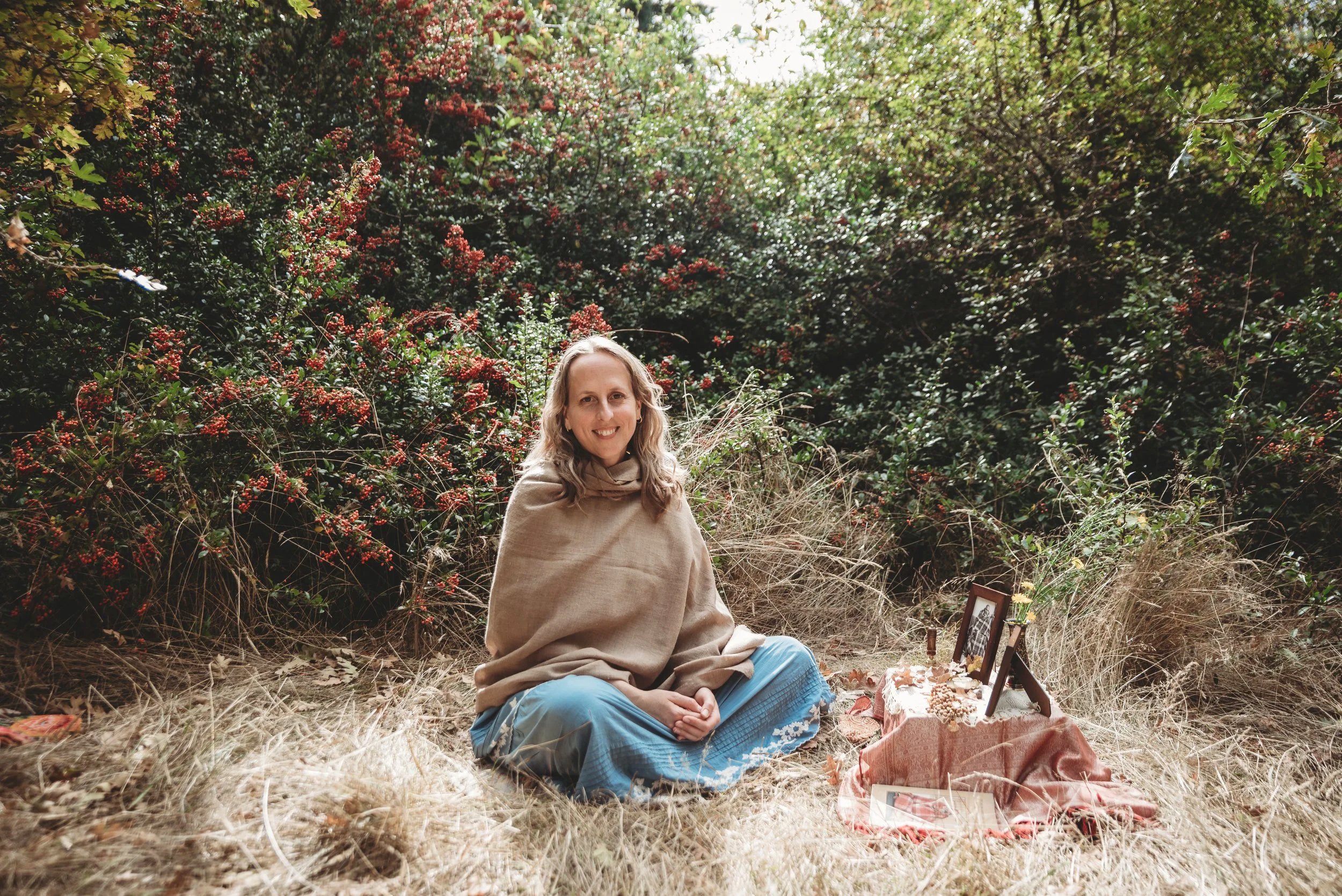 A woman sitting cross-legged on dry grass in a natural setting surrounded by green bushes and red berries, with a small table covered with a pink cloth holding framed photos and small decorative items.