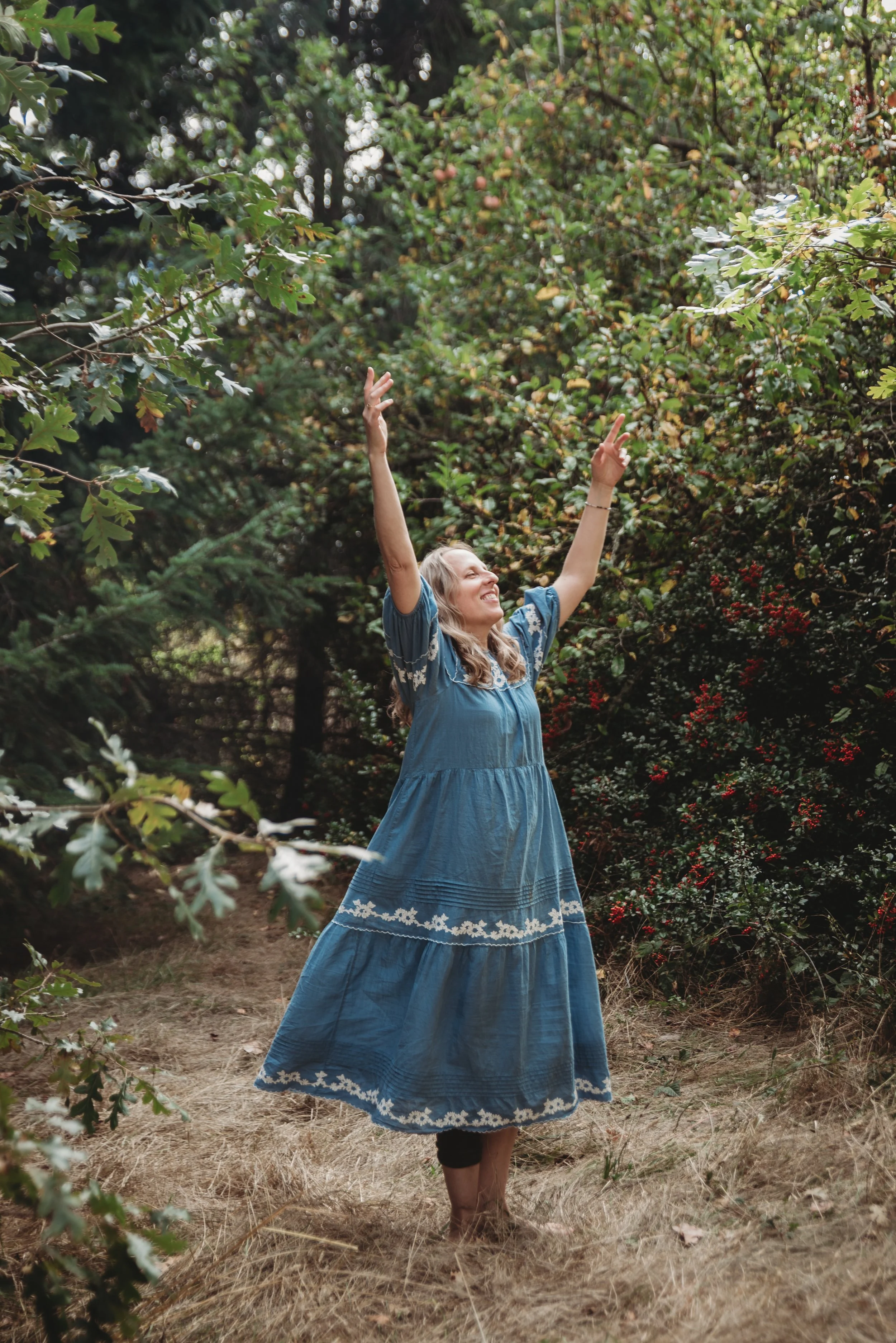A woman with long blonde hair wearing a blue dress with embroidered hem, standing outdoors in a wooded area with leafy trees and bushes, raising her arms with a joyful expression.
