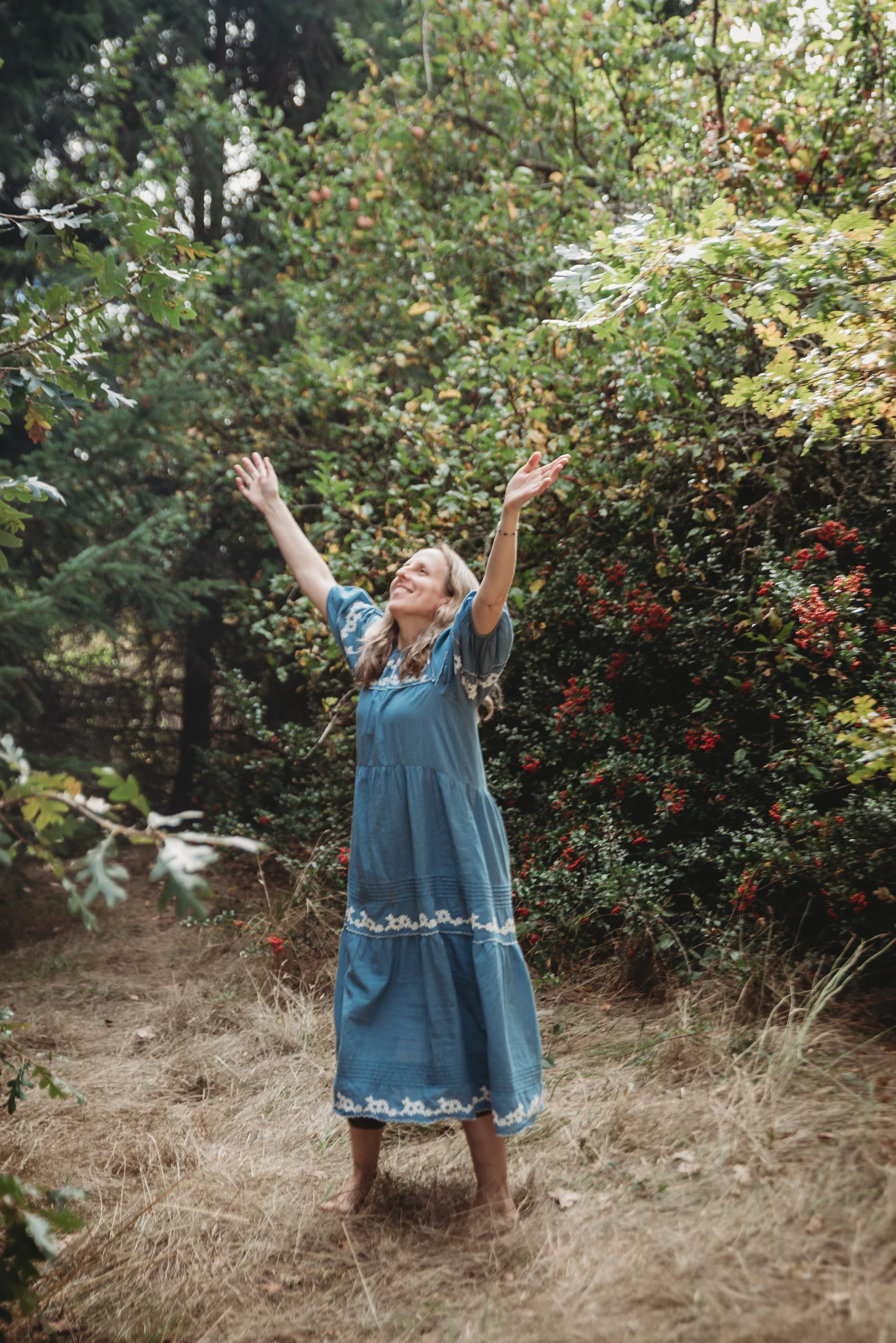 A woman with long blonde hair in a blue dress with white embroidery, standing outdoors on a dry grassy area, raising her arms with a joyful expression. She is surrounded by bushes with green leaves and red berries.