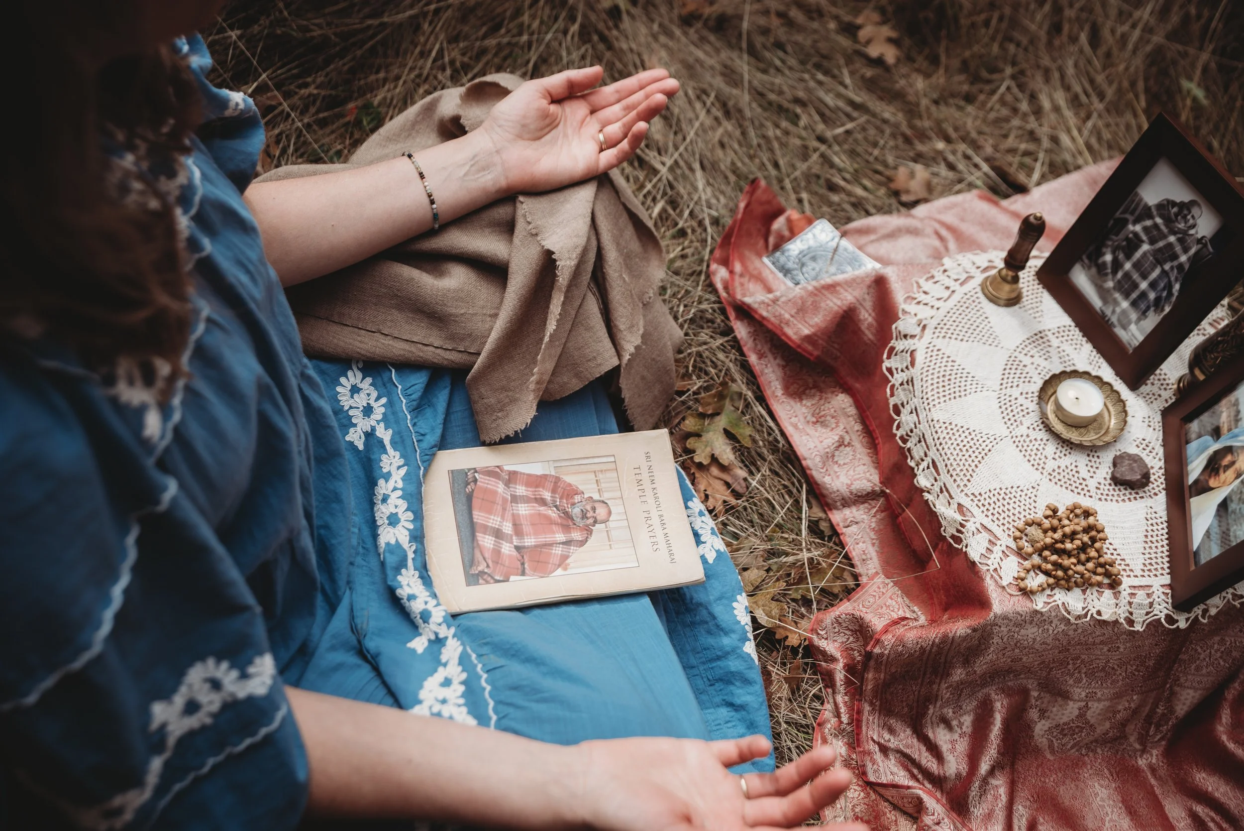 A person sitting on the ground with hands raised in prayer, wearing a blue embroidered dress and a beige shawl, nearby a small altar with framed photos, candles, a Bible, and spiritual objects on a red and white cloth.