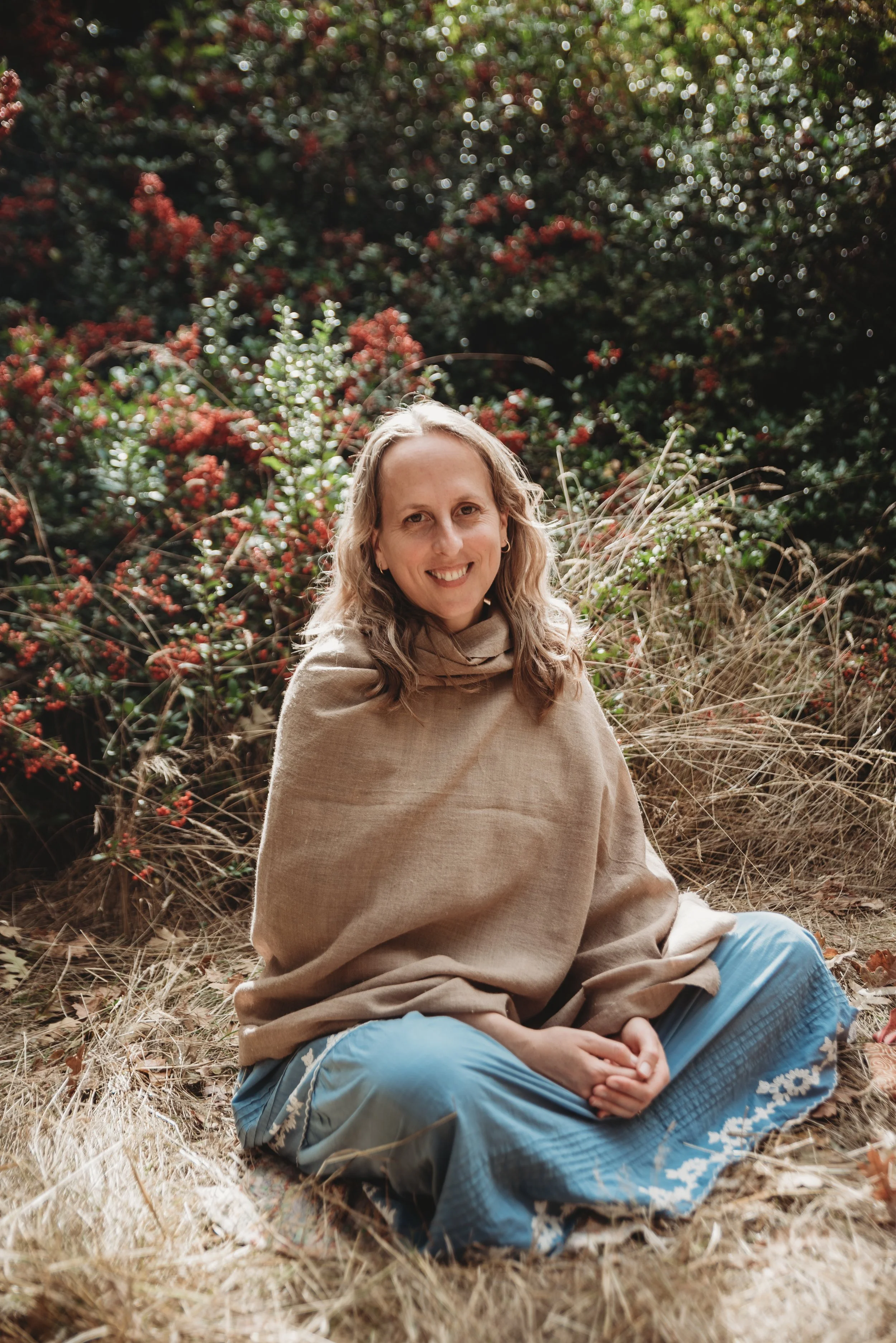 A woman sitting outdoors on dry grass, smiling at the camera, with green and red foliage in the background.