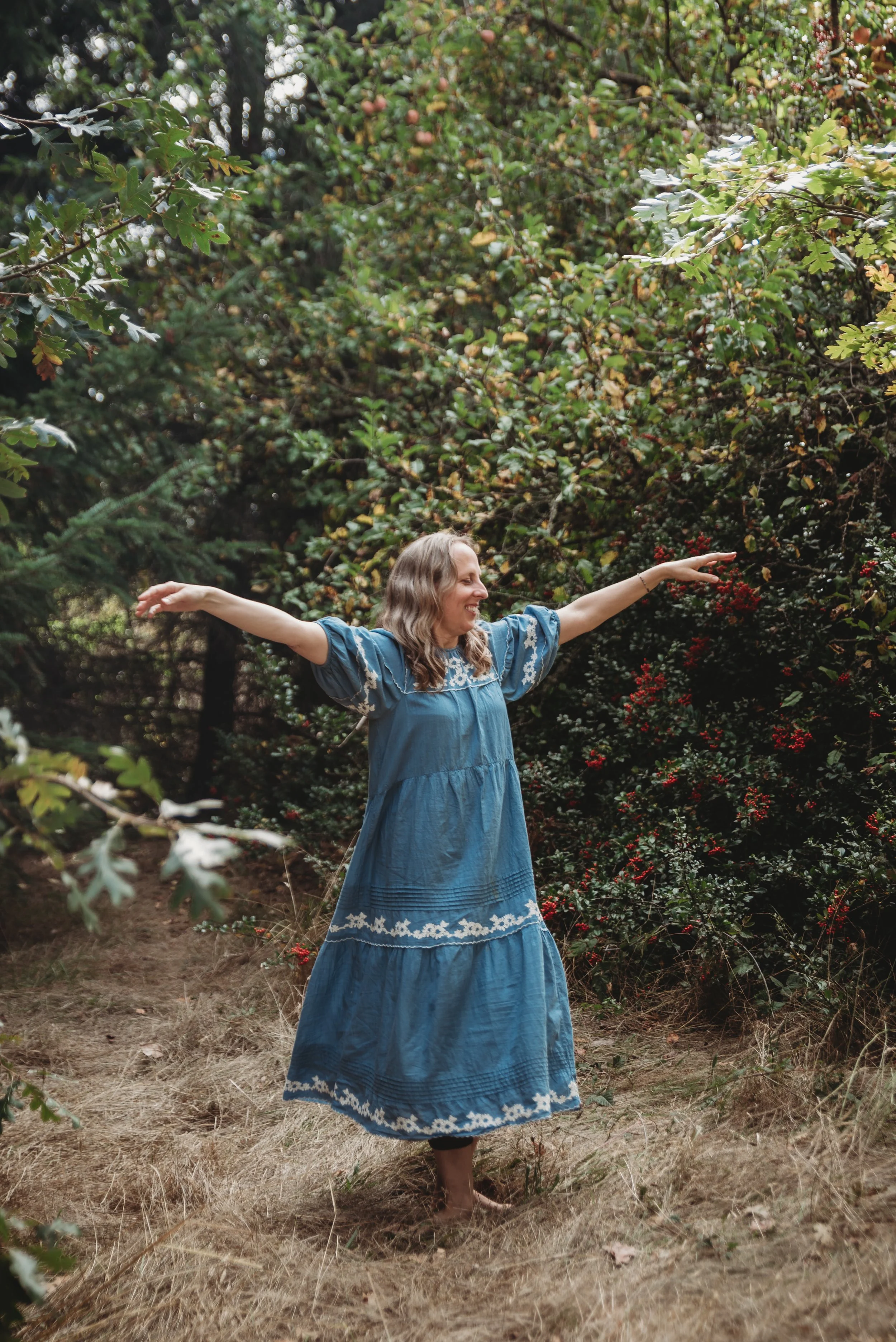 A woman in a blue dress dancing barefoot in a forest clearing with her arms outstretched and surrounded by green foliage and red berries.