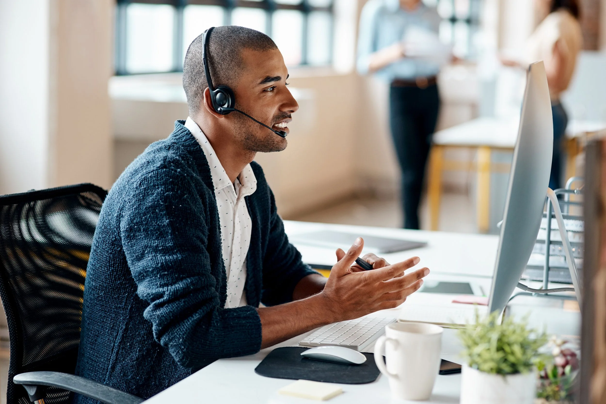 A man wearing a headset and a blue sweater sitting at a desk in an office, working on a computer with a mouse and keyboard, with blurred people working in the background.