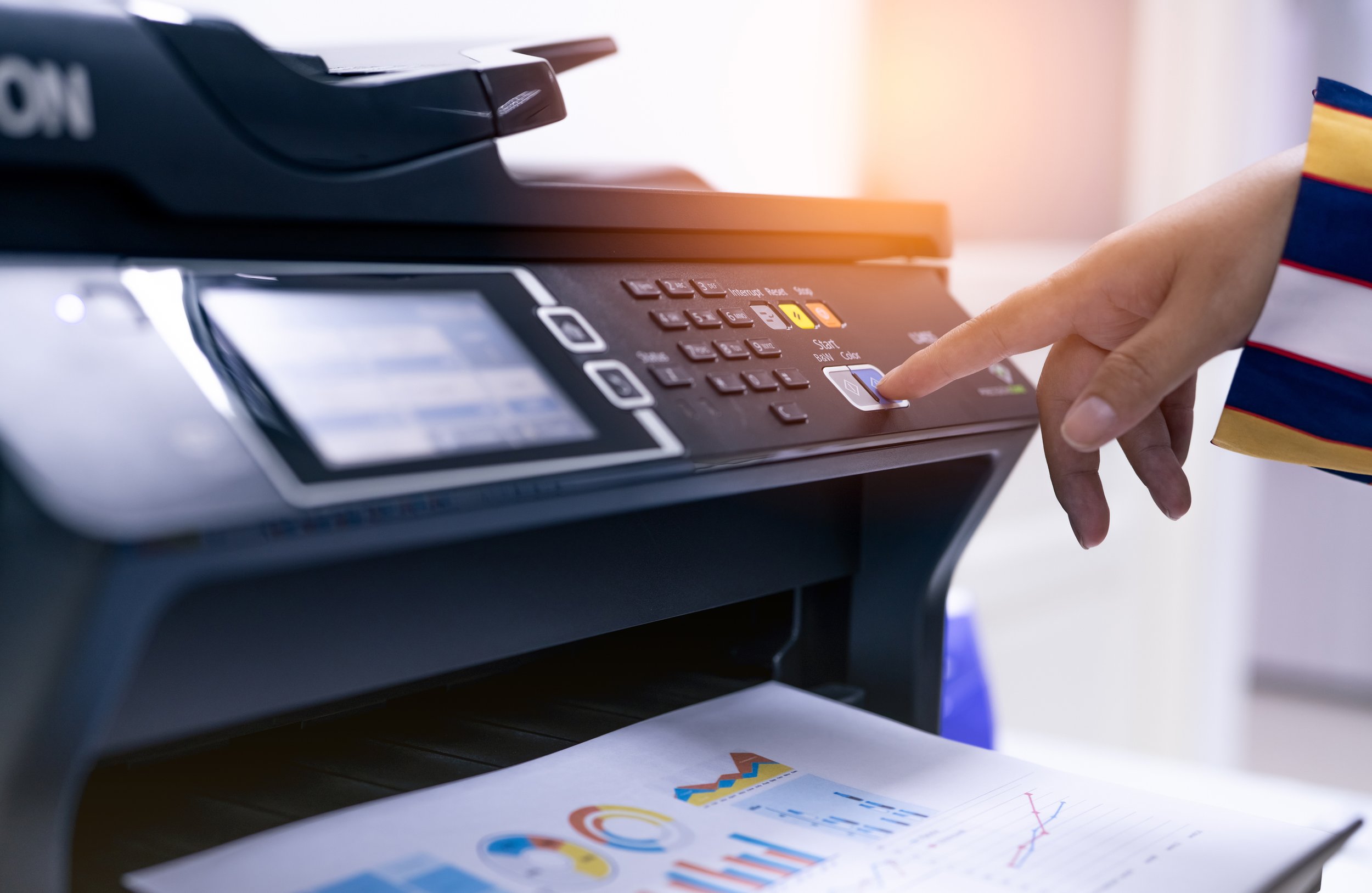 A person's hand pressing a button on a black office copier or printer with colorful charts and graphs printed on paper underneath.