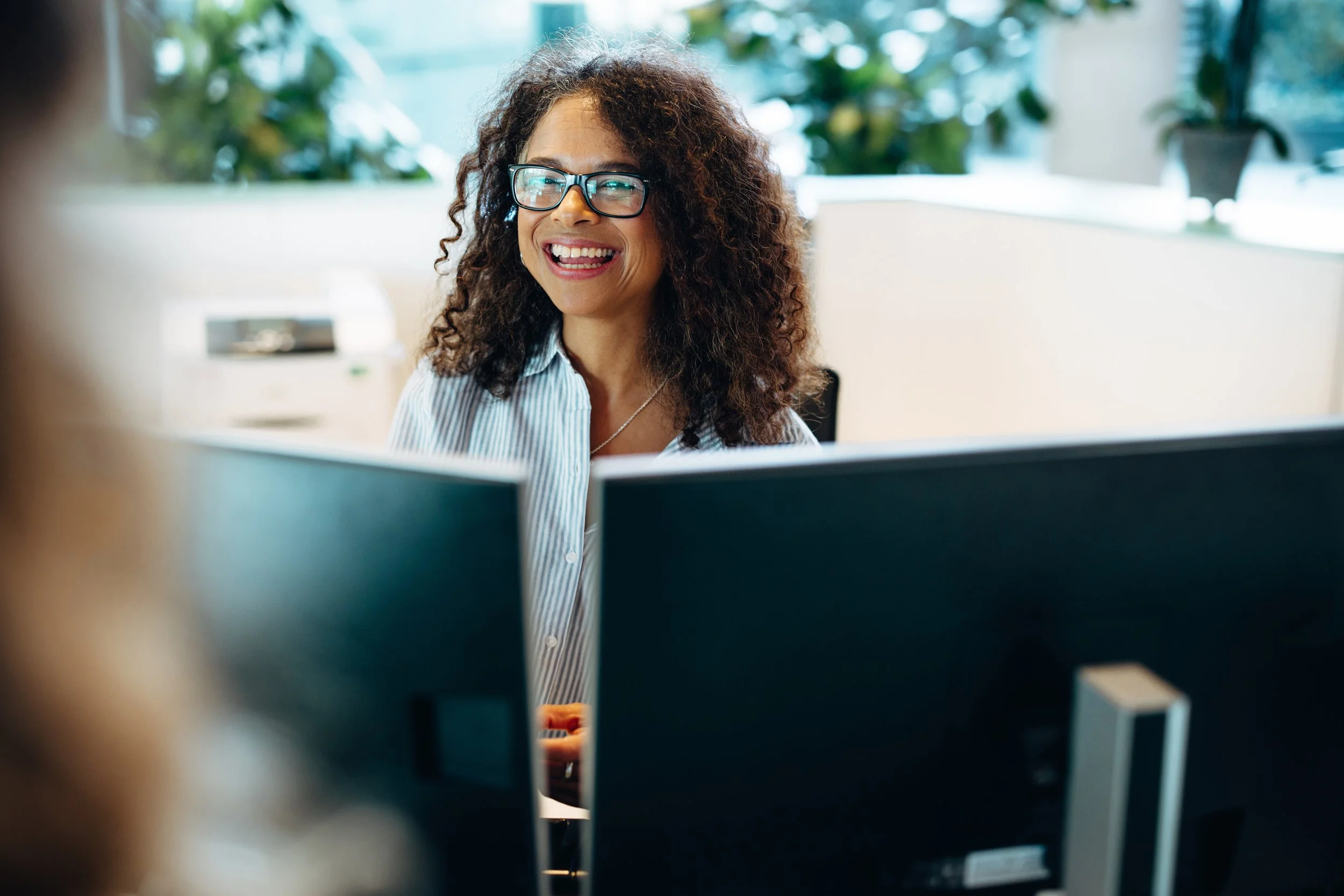 A woman with curly hair and glasses smiling at her computer screen in an office setting.