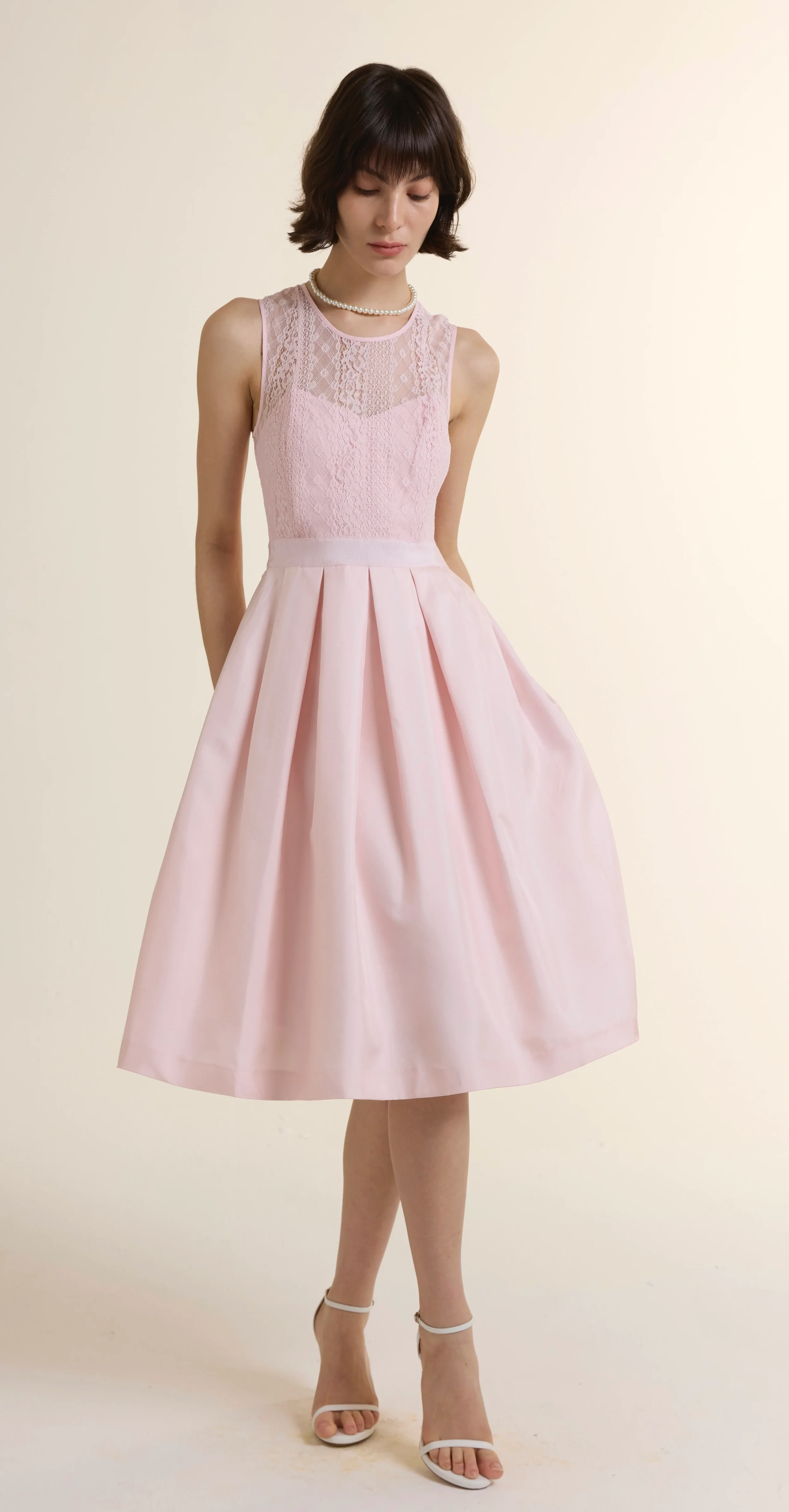 Full-length shot of a model wearing a pale pink sleeveless gown featuring an intricate lace bodice and a flowing 100% silk midi skirt. The elegant design includes a cinched waist and a classic round neckline, styled with white strappy sandals.