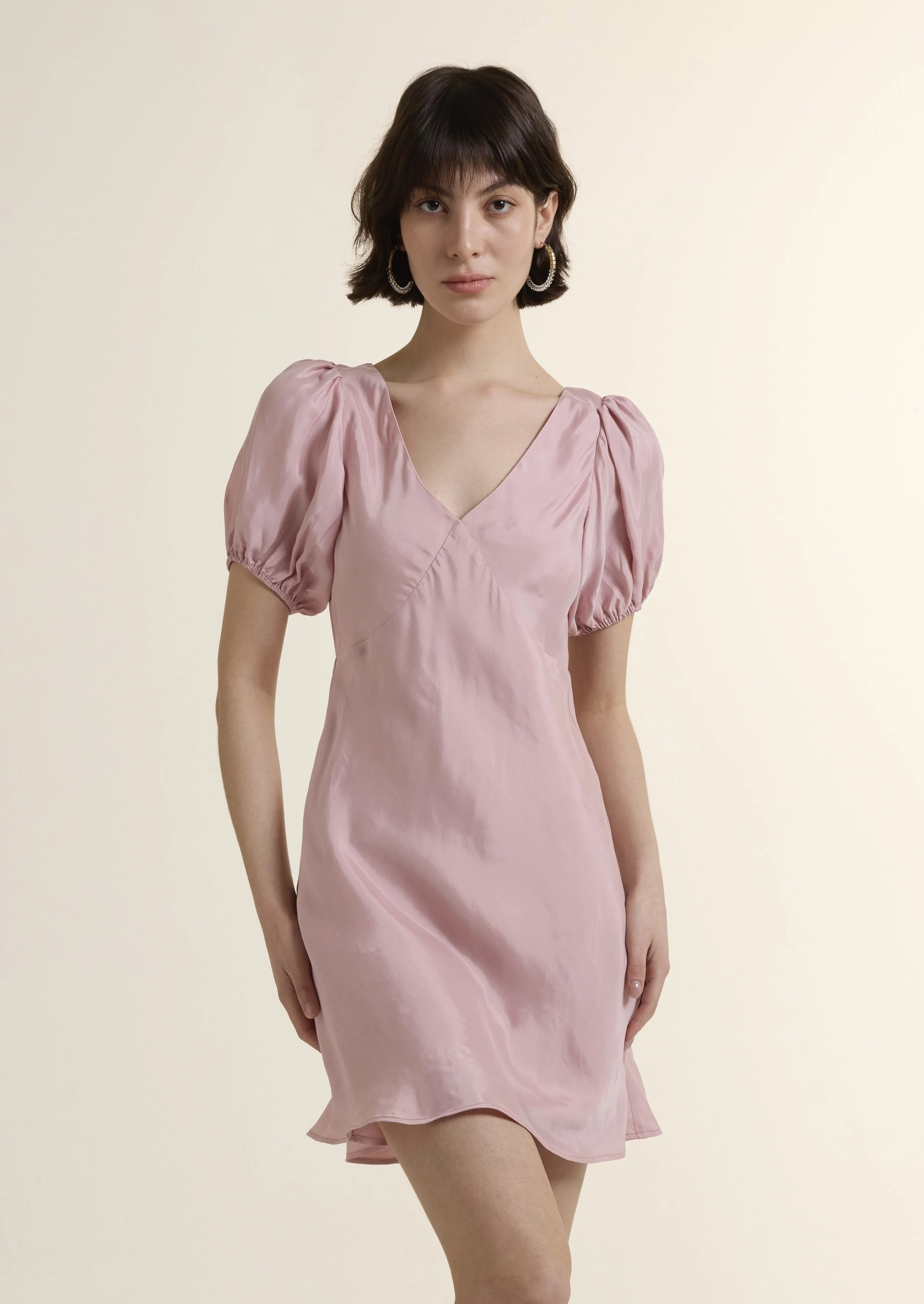 Full-length photo of a model wearing the French Fluid Short Sleeves Mini Dress in a soft dusty pink. The dress features a V-neckline, gathered puff sleeves, and a flowing A-line silhouette, crafted from a luxurious Cupro-Viscose blend.