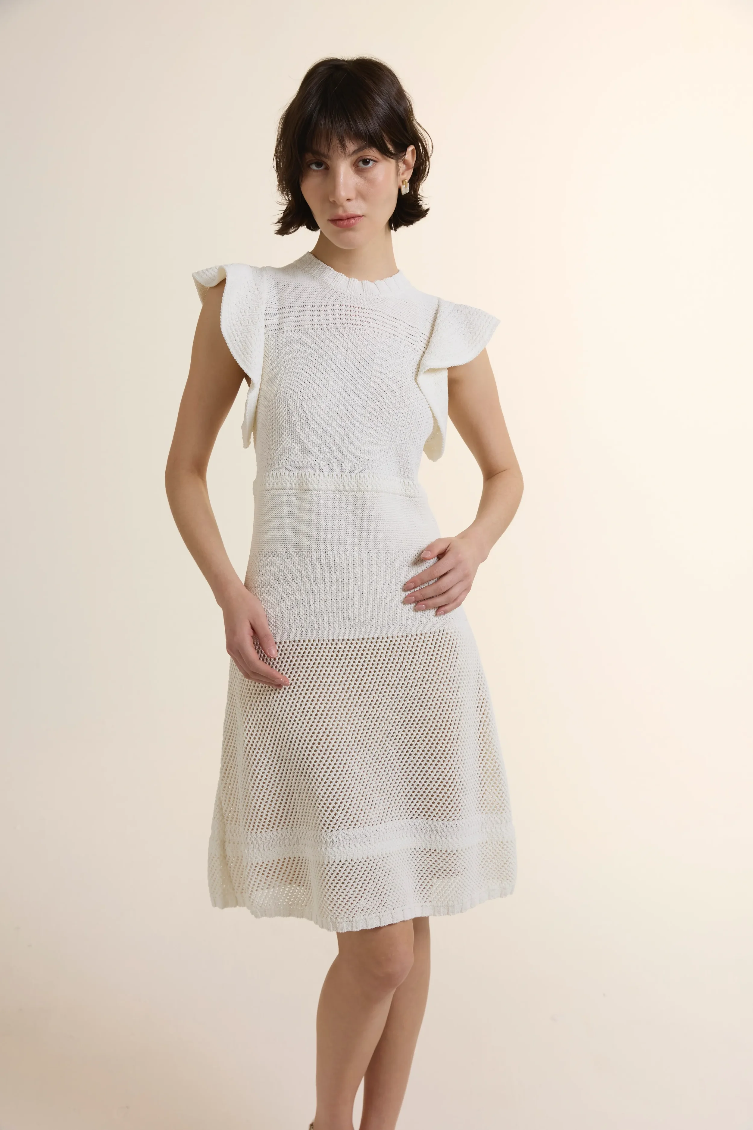A model wearing a white sleeveless pointelle knit dress with delicate open-work patterns and feminine ruffle shoulder details. The dress features a structured knit bodice and a semi-sheer flared hem, crafted from a breathable cotton blend.