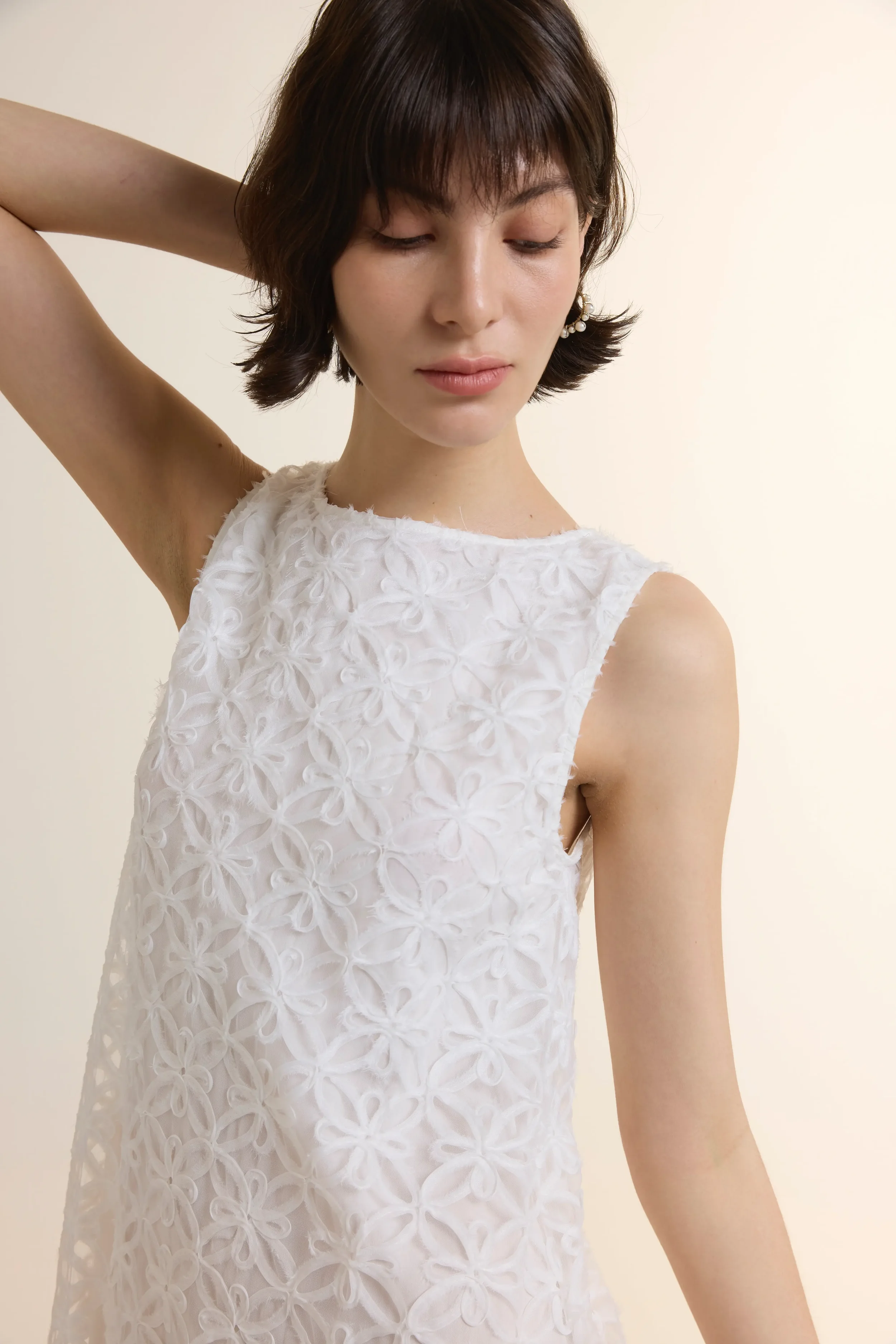 Close-up of Full-length front view of a model wearing a white 3D Floral Midi Dress with an intricate geometric flower lace pattern. The sleeveless dress features a sheer tiered hem and a clean jewel neckline, styled with white strappy heels.