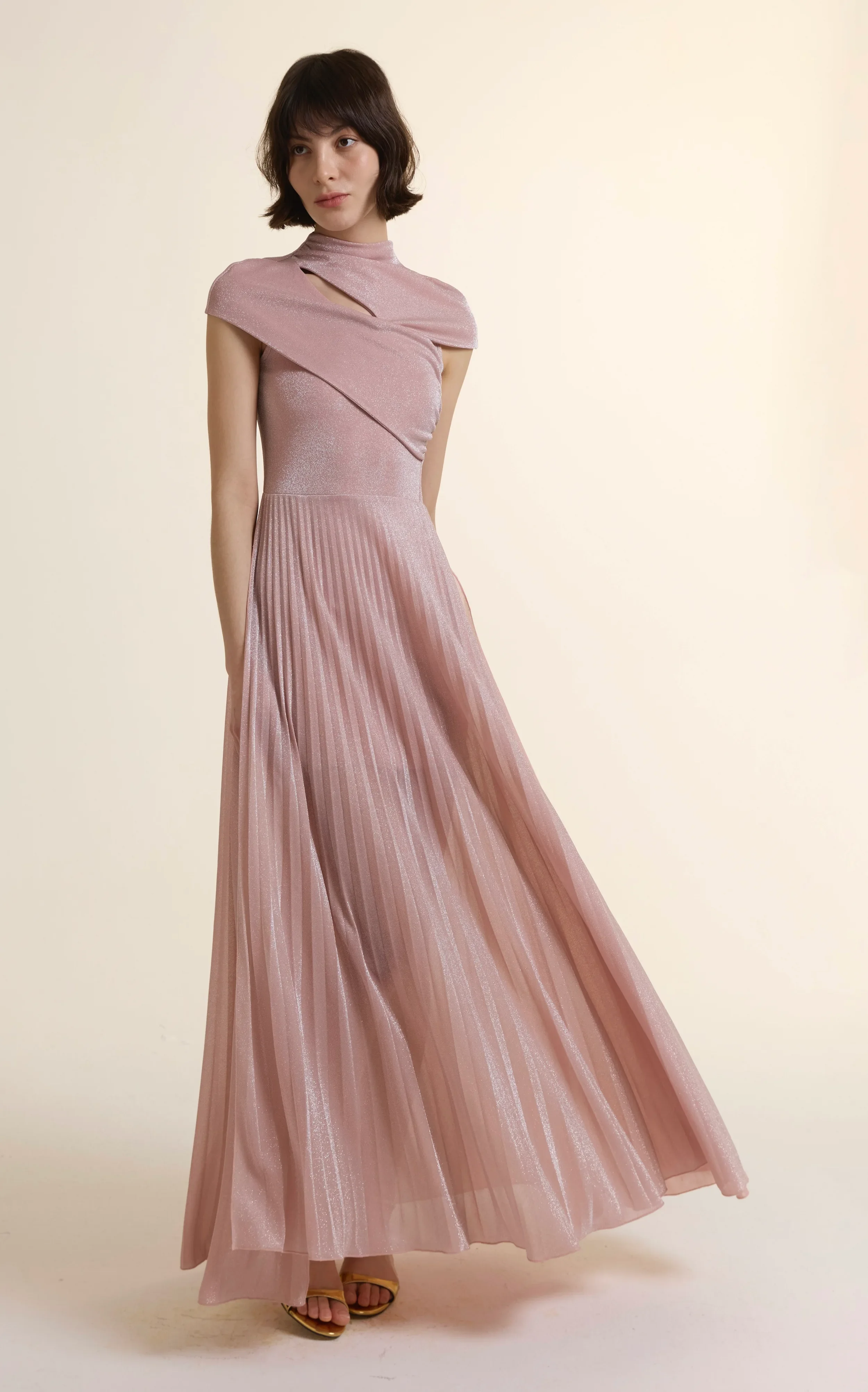 Full-length view of a model wearing a shimmering dusty pink Luminous Pleated Maxi Gown. The dress features a mock neckline with a sculptural crossover bodice and a high-waisted, finely pleated maxi skirt that creates a fluid, elongated silhouette.