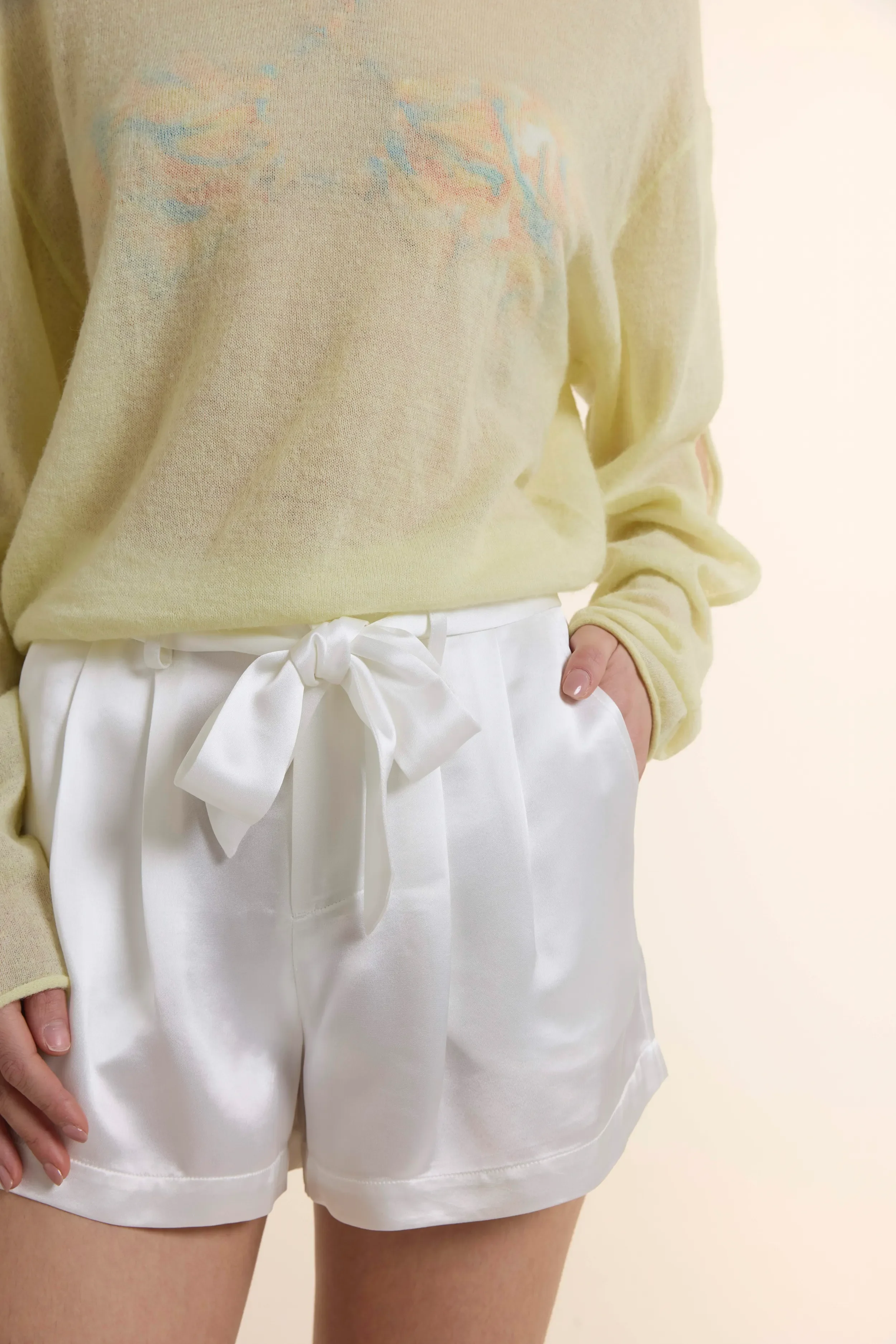 Close-up of white Satin Loose Shorts featuring a high-waisted fit with a sophisticated bow-tie belt. The fabric showcases a luxurious luster from the acetate-polyester blend, styled with a hand in the side pocket.