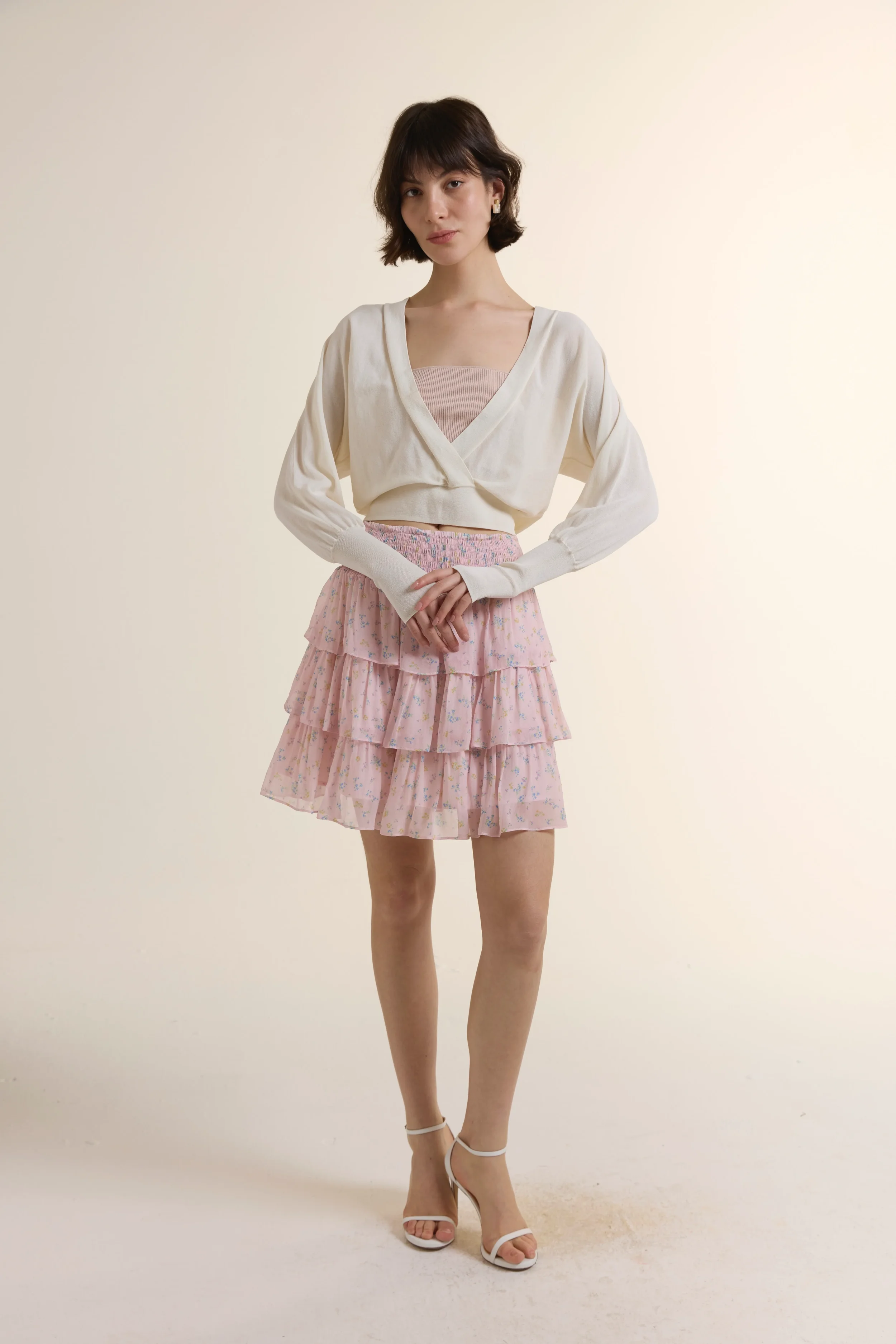 A full shot of a model wearing a cream-colored lightweight wrap knit top paired with a pink floral georgette layered mini skirt. The look highlights the soft texture of the knit and the fluid, tiered ruffles of the floral skirt.