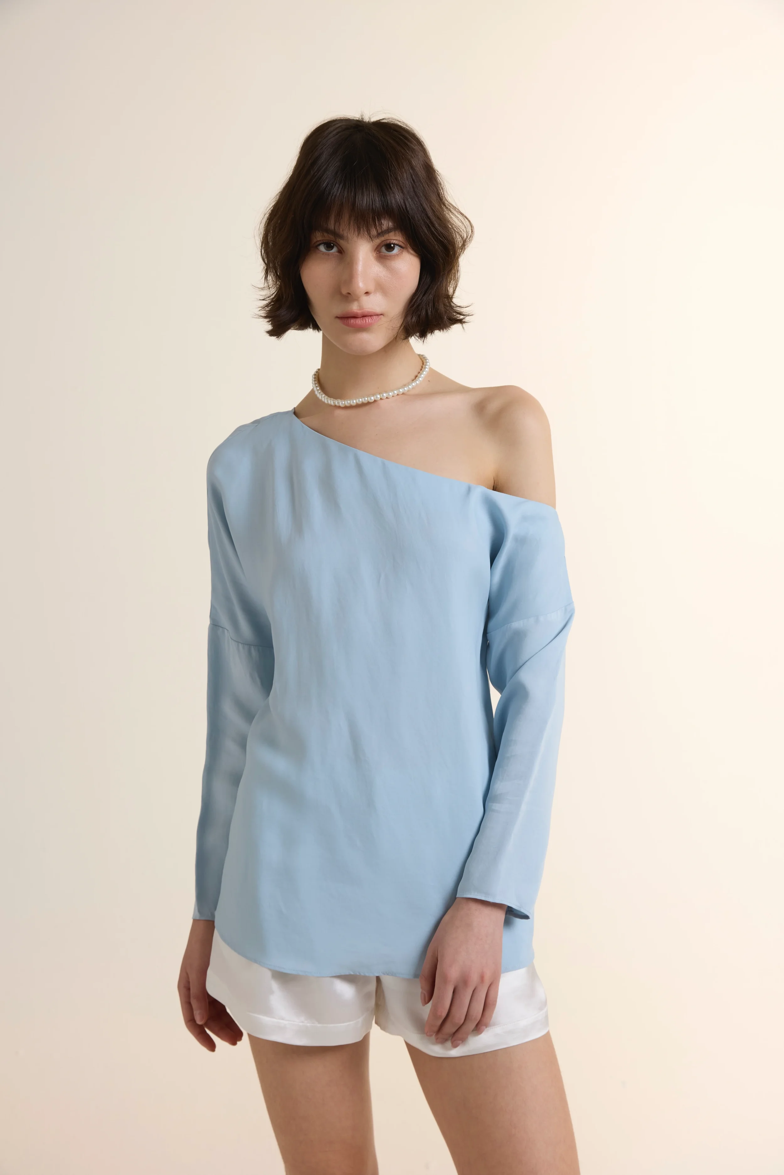 Front view of a woman wearing a light blue one-shoulder long-sleeve top made of Tencel and Viscose, paired with white silk shorts, against a minimalist cream background.