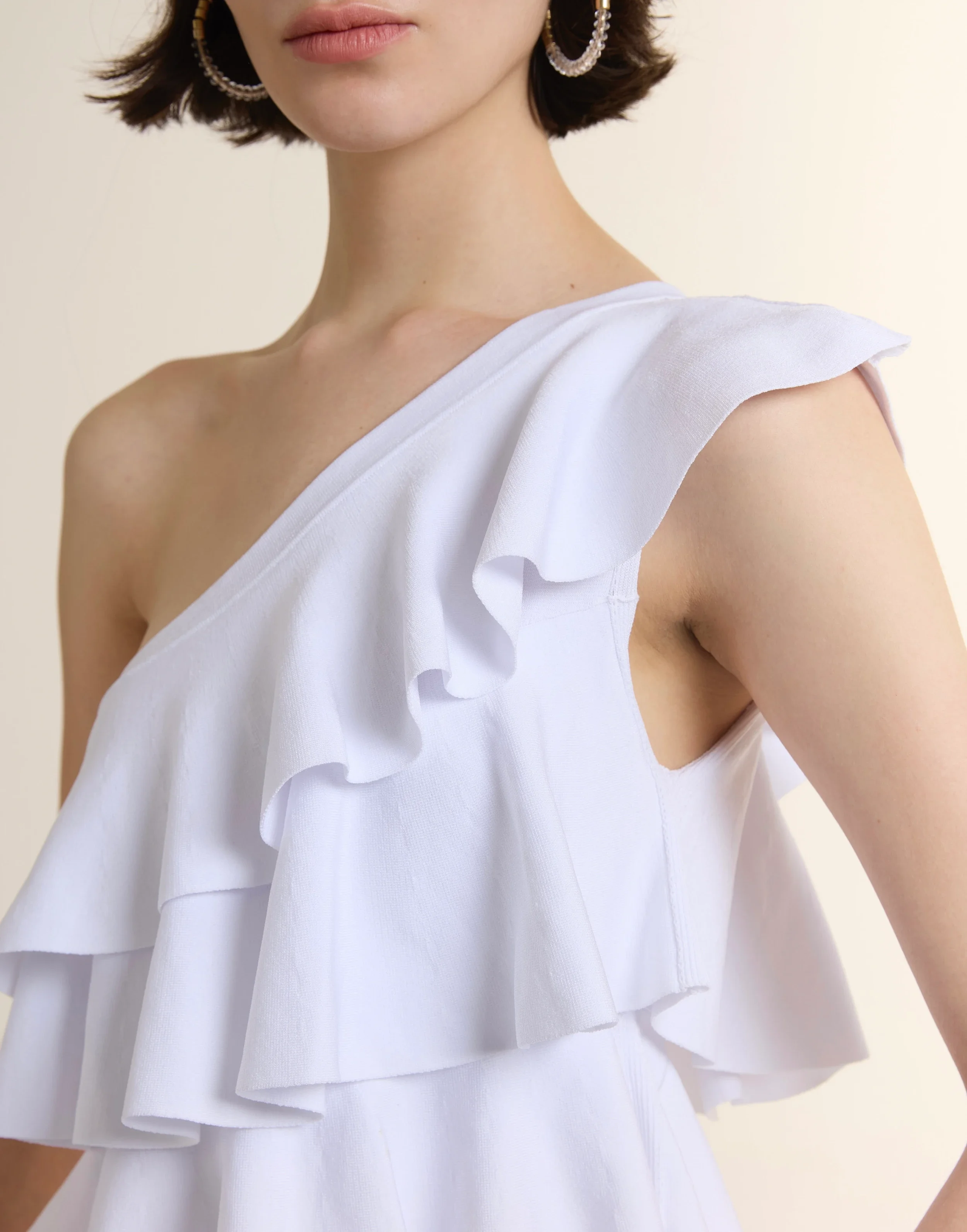 Close-up of a model wearing a white sculpted one-shoulder knit top with tiered ruffle layers, paired with blue denim jeans. The fabric features a fine ribbed texture in a high-elastic viscose blend.