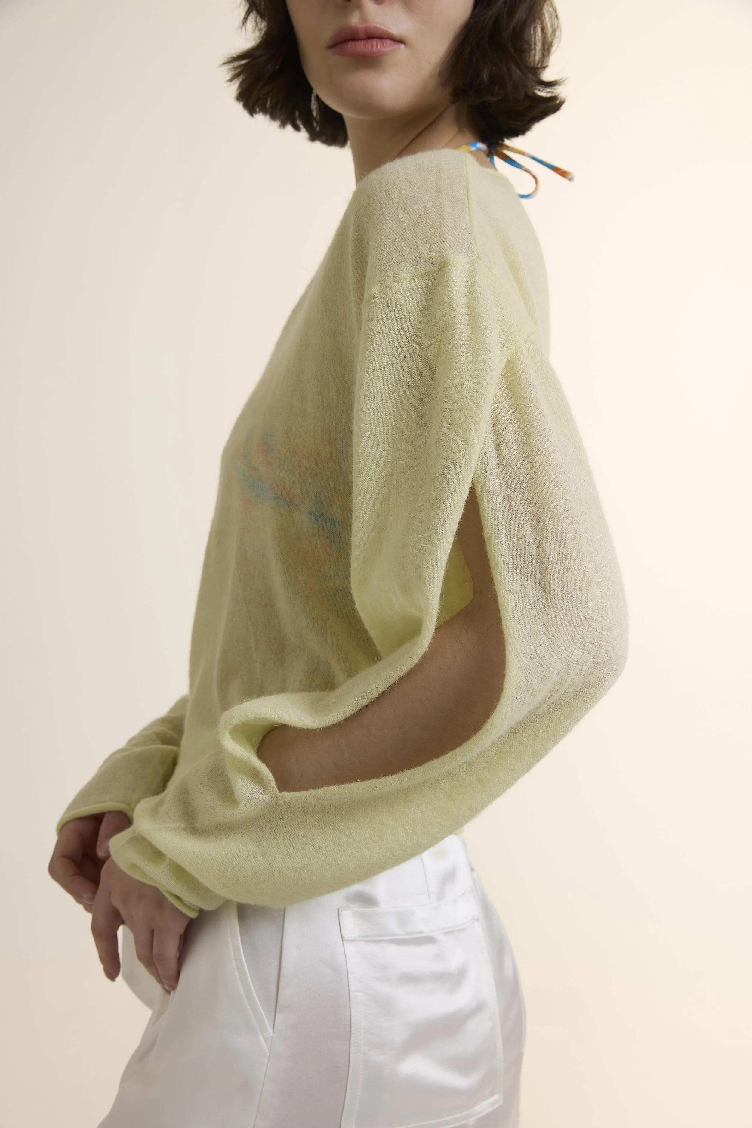 A model wearing a light lime-green sheer knit sweater over a patterned bikini top, paired with white high-waisted satin loose shorts. The look highlights the delicate alpaca-wool blend texture and the elegant satin sheen of the shorts.