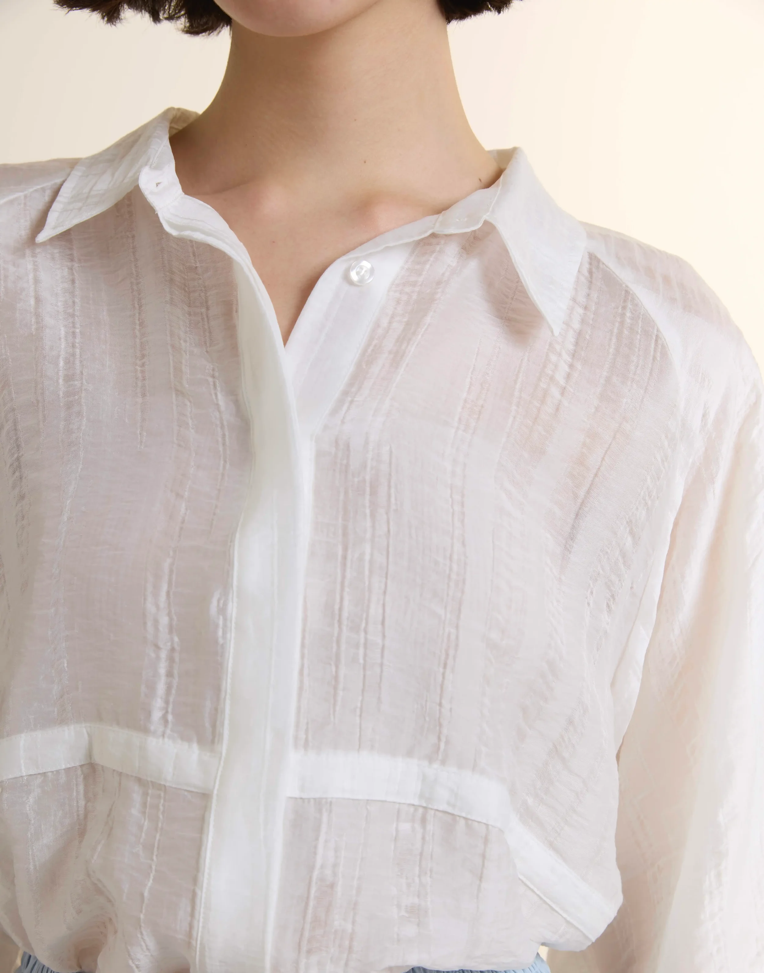 Close-up of a white sheer oversized button-down shirt made from a breathable Tencel and Nylon blend. Features dropped shoulders, a classic collar, and a subtle grid-like texture for a contemporary, light luxury feel.