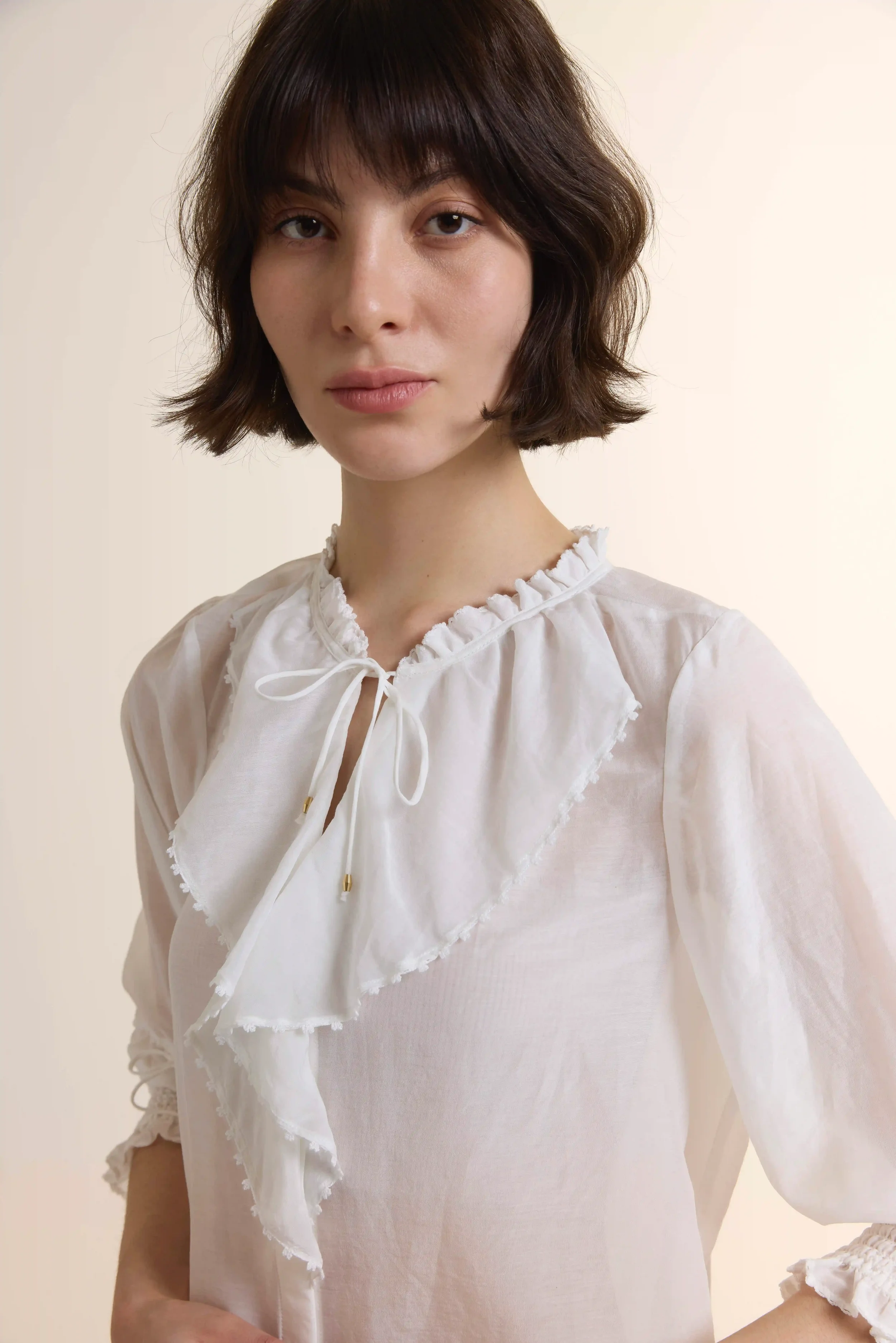 A close-up view of the Ruffle Cotton-Silk Blouse, showcasing the delicate ruffled neckline with tie-up detailing and the subtle sheer luster of the 70% cotton and 30% silk fabric blend.