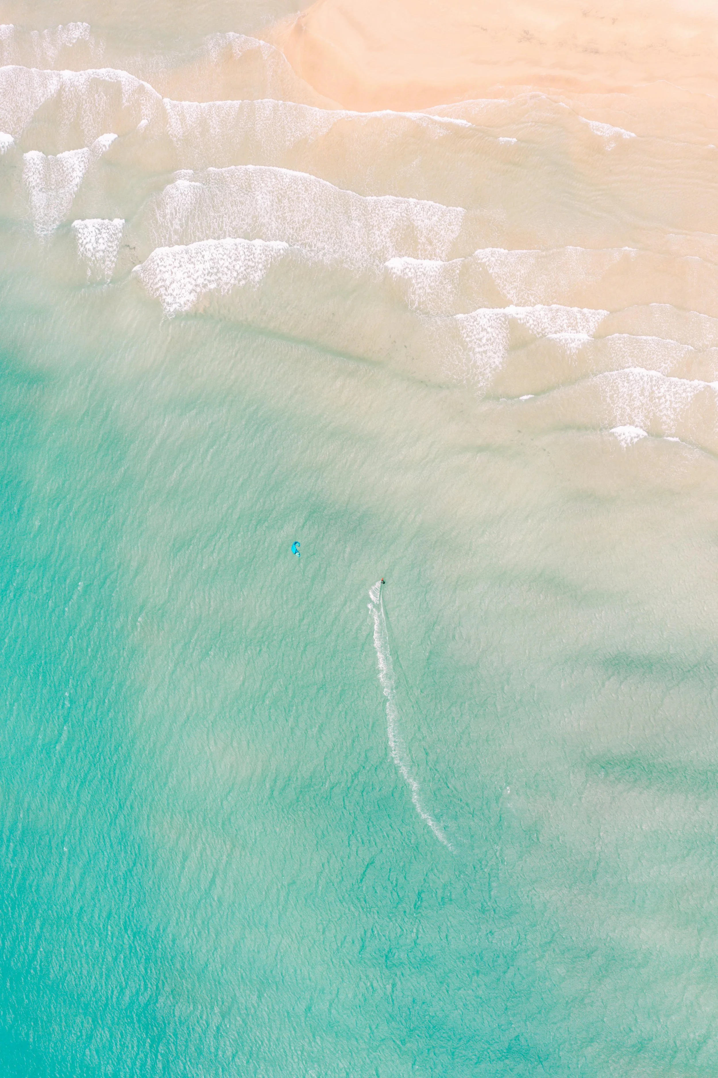 Aerial view of beaches with waves crashing onto the shore and people in the water, featuring clear turquoise and sandy areas.