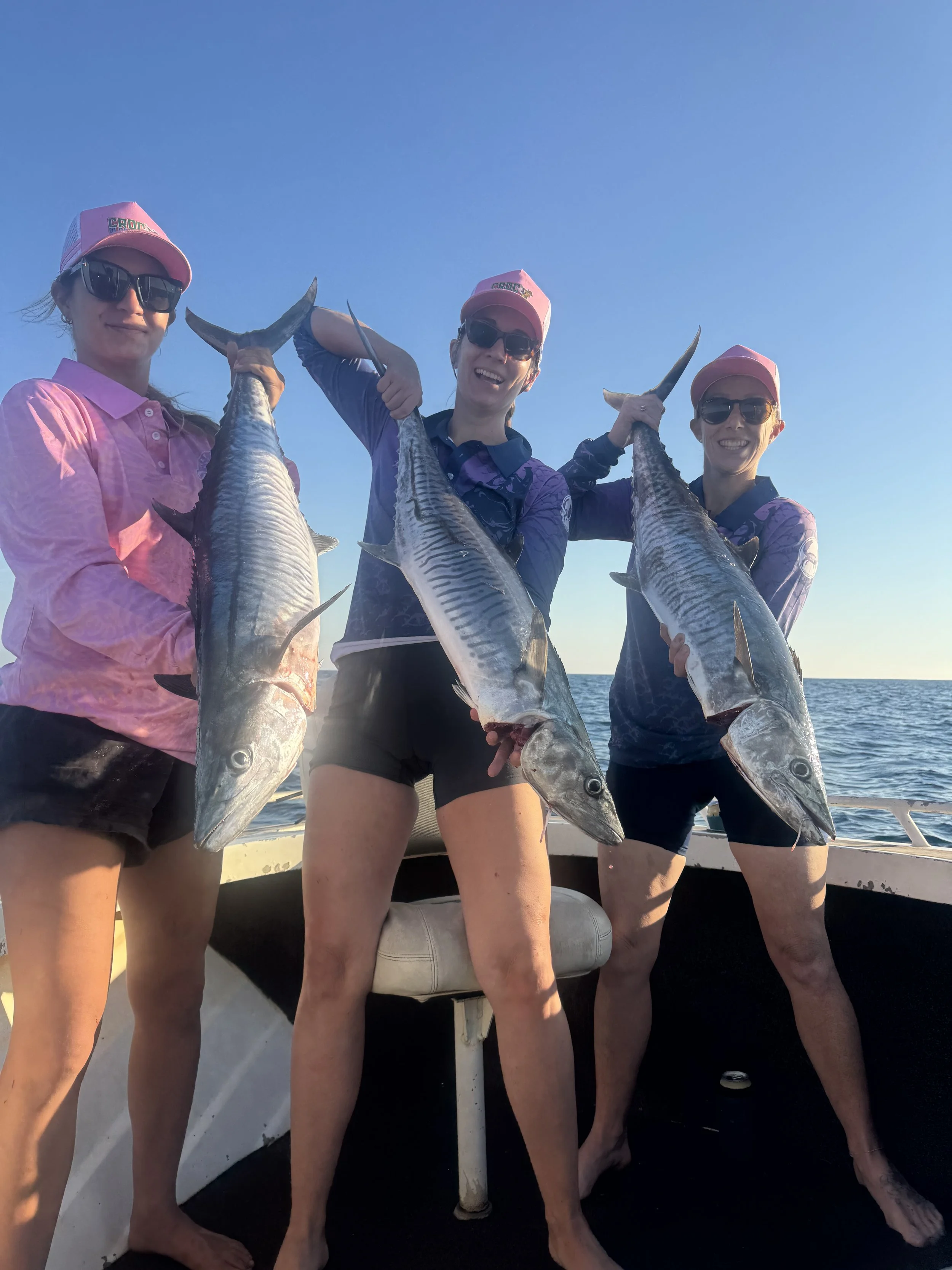 Three women on a boat holding up large fish they caught, with a clear blue sky and ocean in the background.