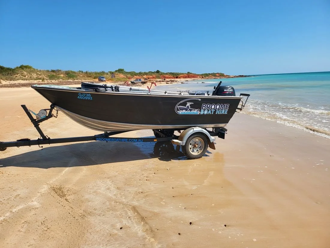 A black boat on a trailer at the edge of a sandy beach with turquoise water and a clear blue sky in the background.