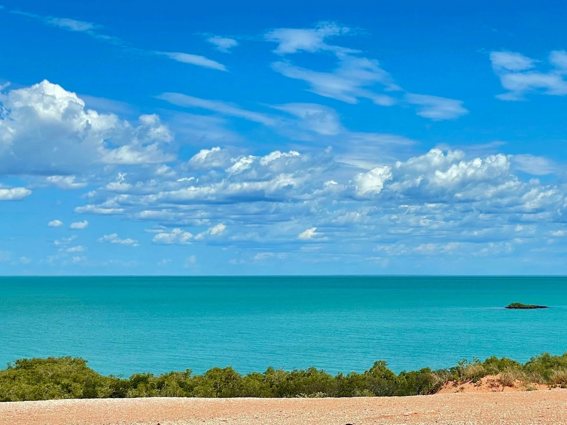 A scenic view of a sandy beach with green bushes in the foreground, a turquoise ocean, and a blue sky with scattered clouds.