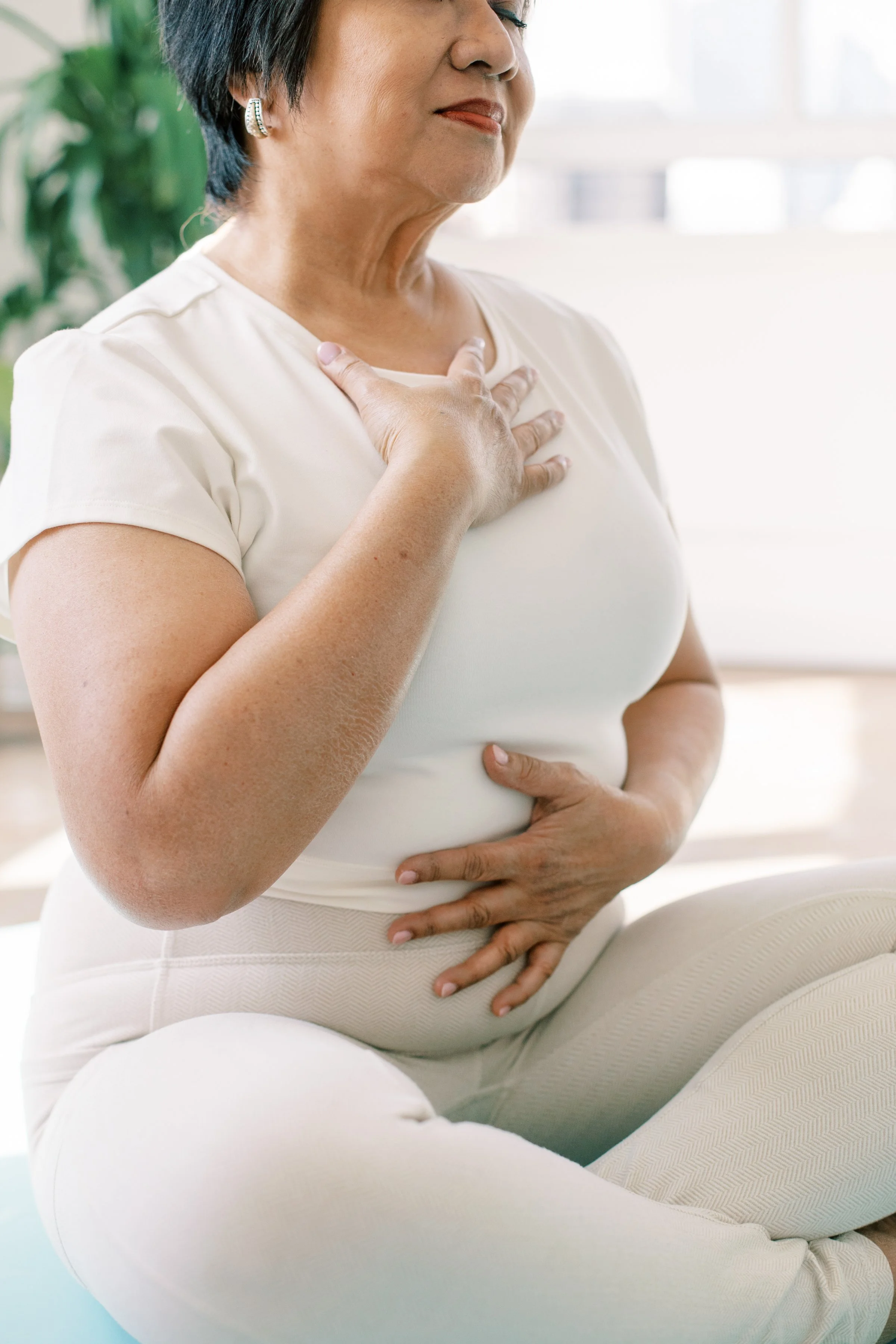 An older woman practicing yoga or meditation indoors, sitting cross-legged with her hand on her chest and stomach, doing a self-heart and stomach center focus.