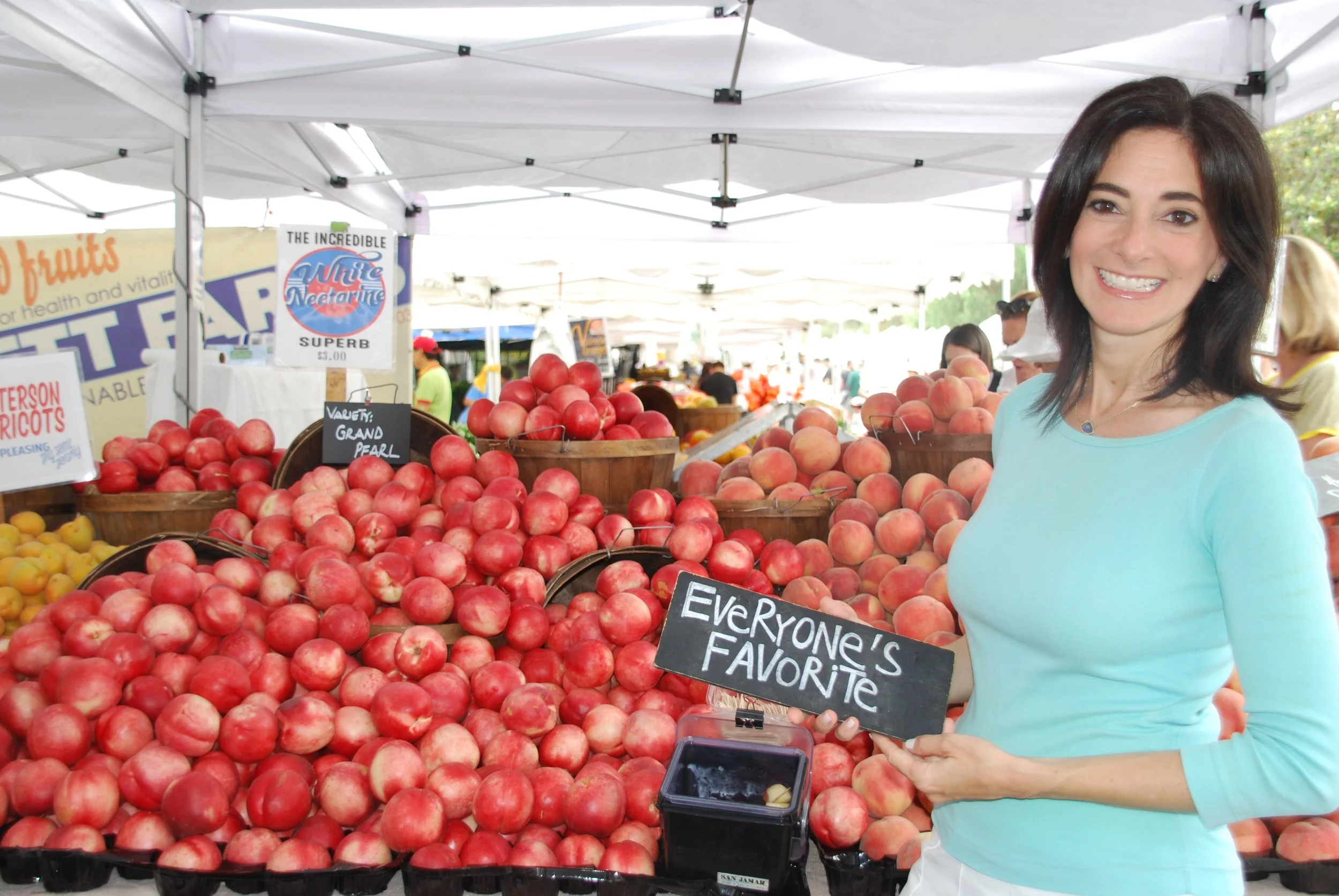 A woman at a farmer's market holding a chalkboard sign that reads 'Everyone's Favorite', with baskets of red and pink apples around her under a white canopy, and other market stalls and shoppers in the background.