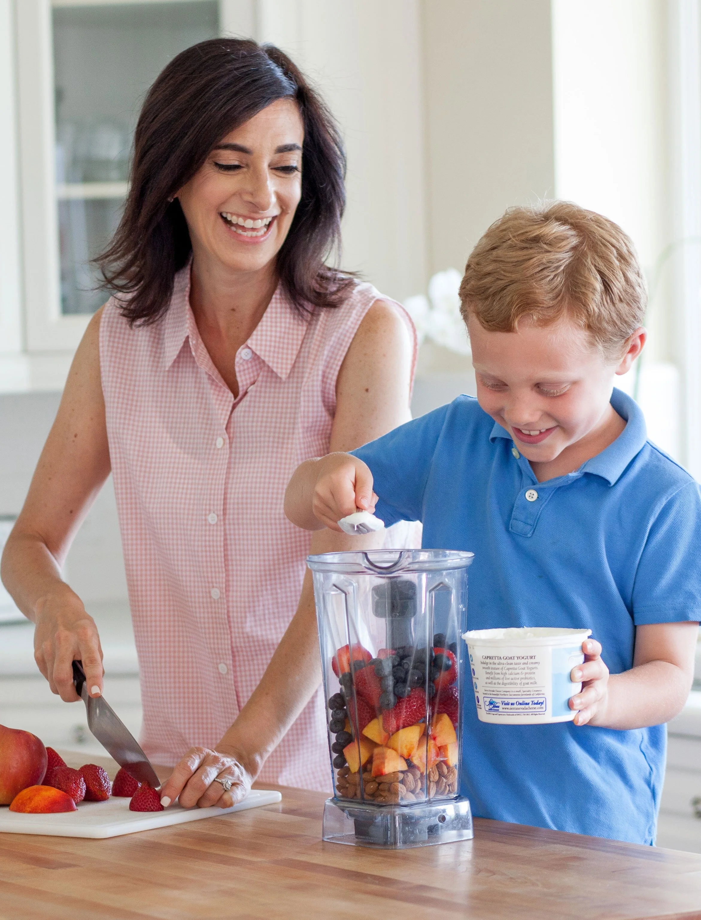 A woman and a boy in a kitchen preparing a smoothie with fresh fruit and yogurt.