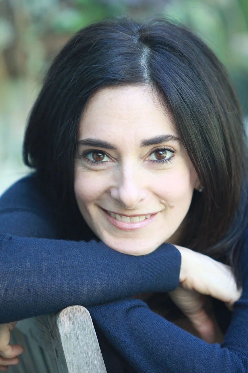 A woman with dark hair smiling, resting her chin on her arm, outdoors with a blurred natural background.