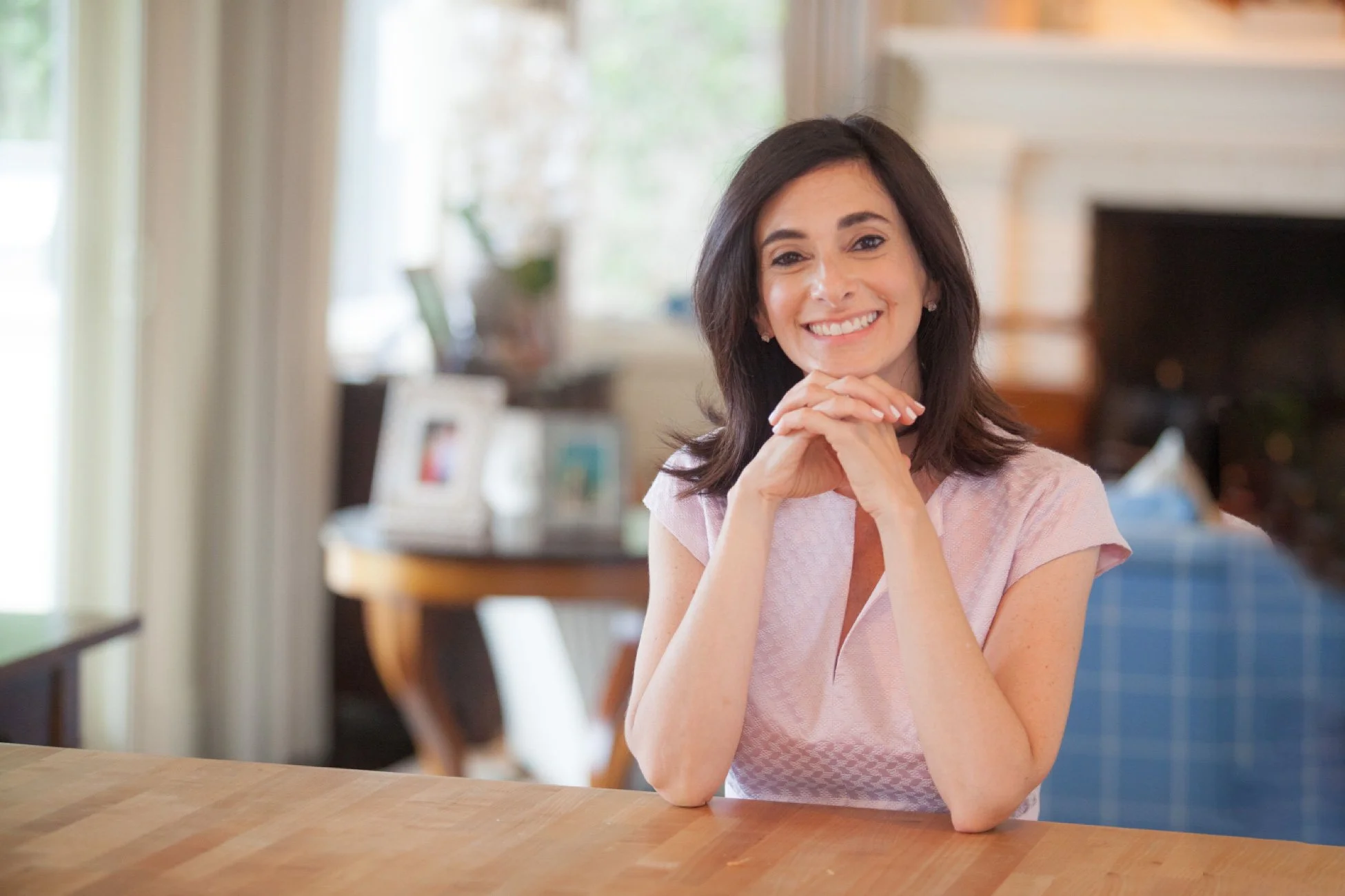 A smiling woman with dark hair, wearing a light pink top, sitting at a wooden table in a cozy, well-lit living room with a fireplace and family photos.