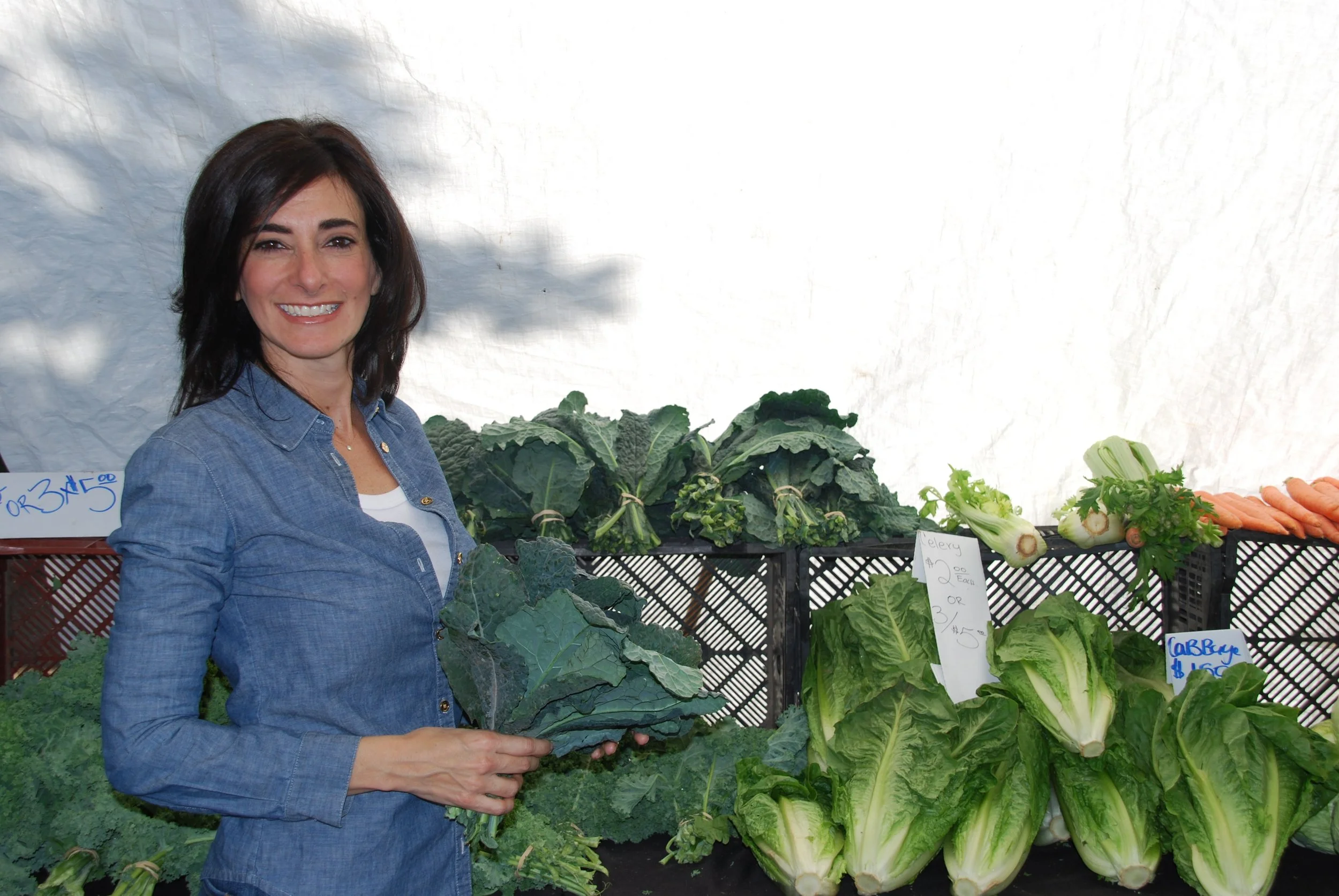 Woman shopping for fresh vegetables at a farmers market, holding kale, with other greens and carrots on display.