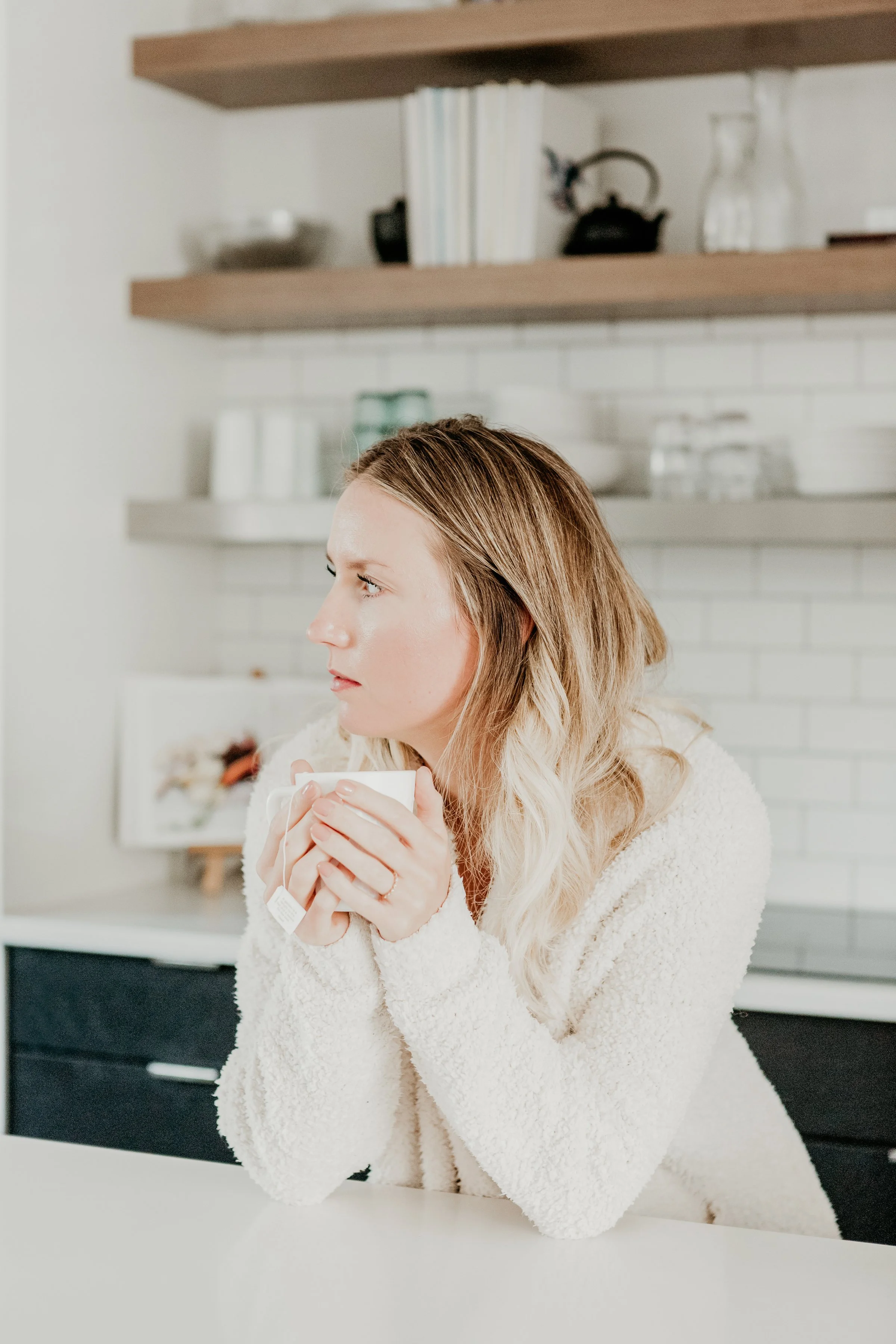 A woman with blonde wavy hair sitting at a white kitchen counter, holding a white mug with both hands, looking to her left.