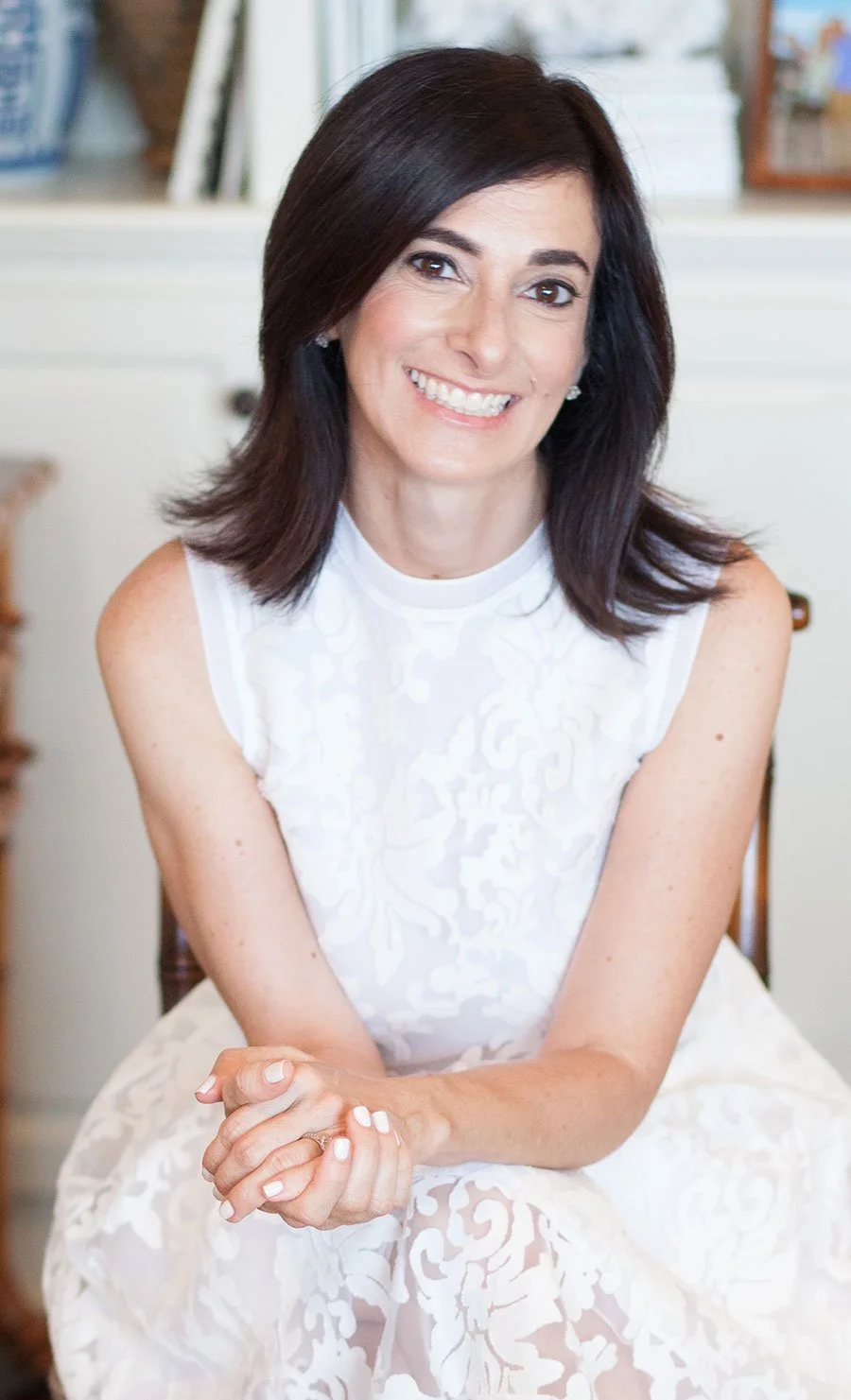 A woman with shoulder-length dark hair smiling, sitting with her hands clasped, wearing a sleeveless white dress with a subtle pattern, in a cozy indoor setting.