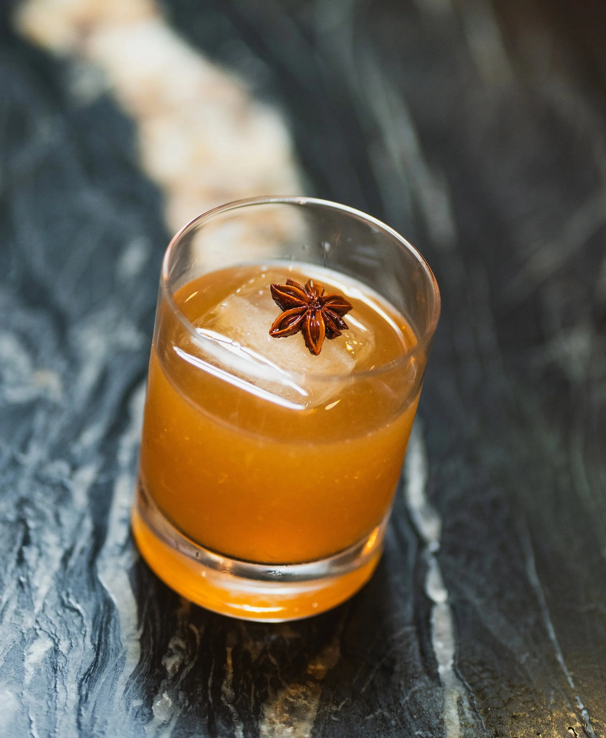 A glass of iced drink garnished with a star anise pod on top, placed on a dark marble bar top.