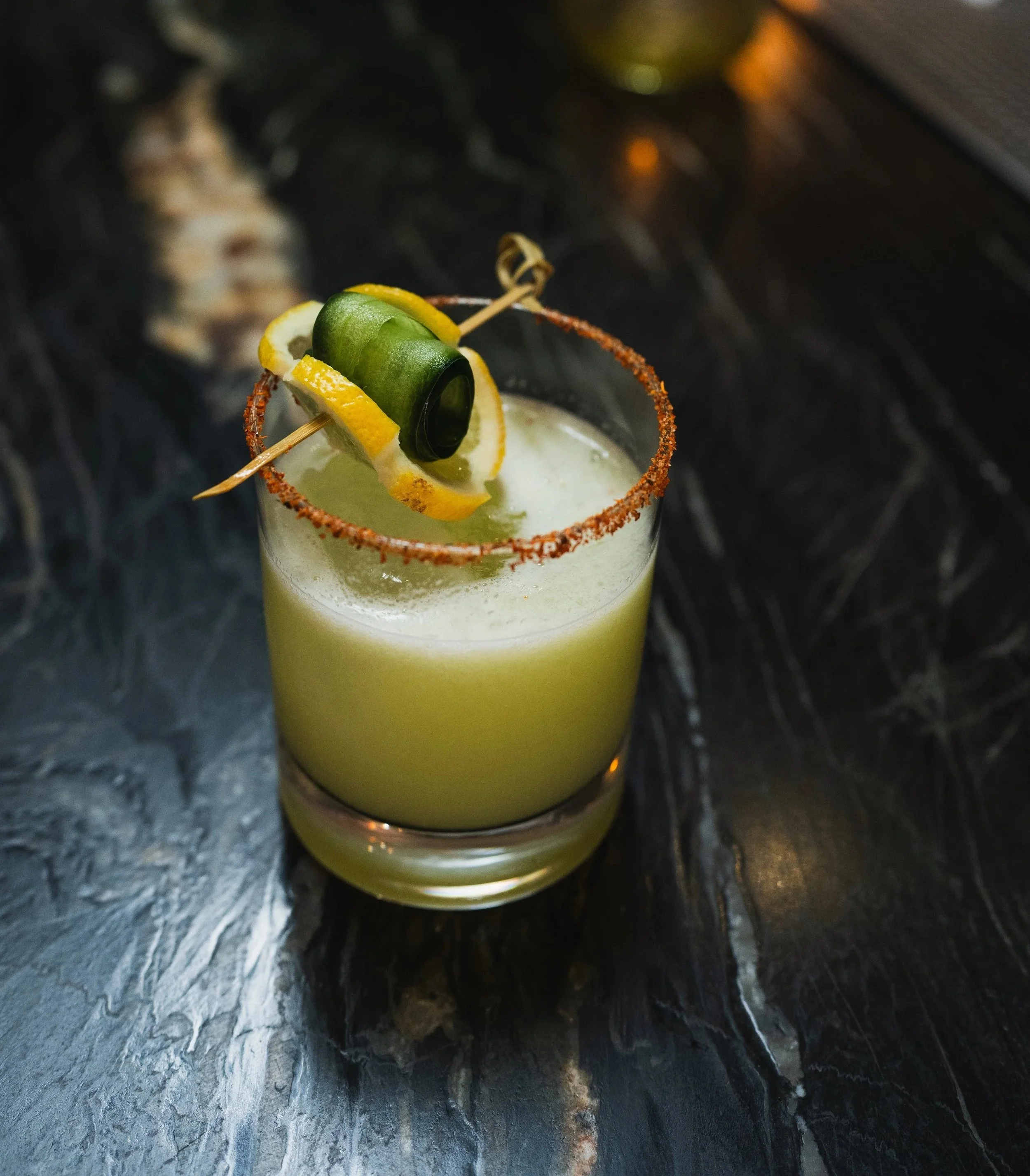 A cocktail in a short glass with a salted rim, garnished with lemon and cucumber on a skewer.