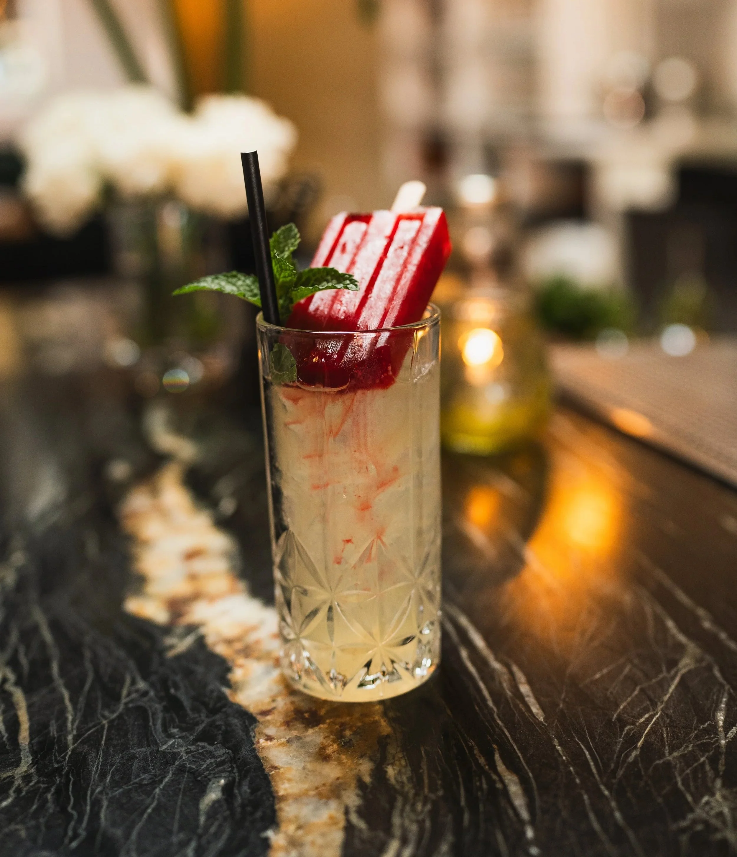 A cocktail in a tall, patterned glass with a red popsicle, a sprig of mint, and black straws on a dark marble bar counter.