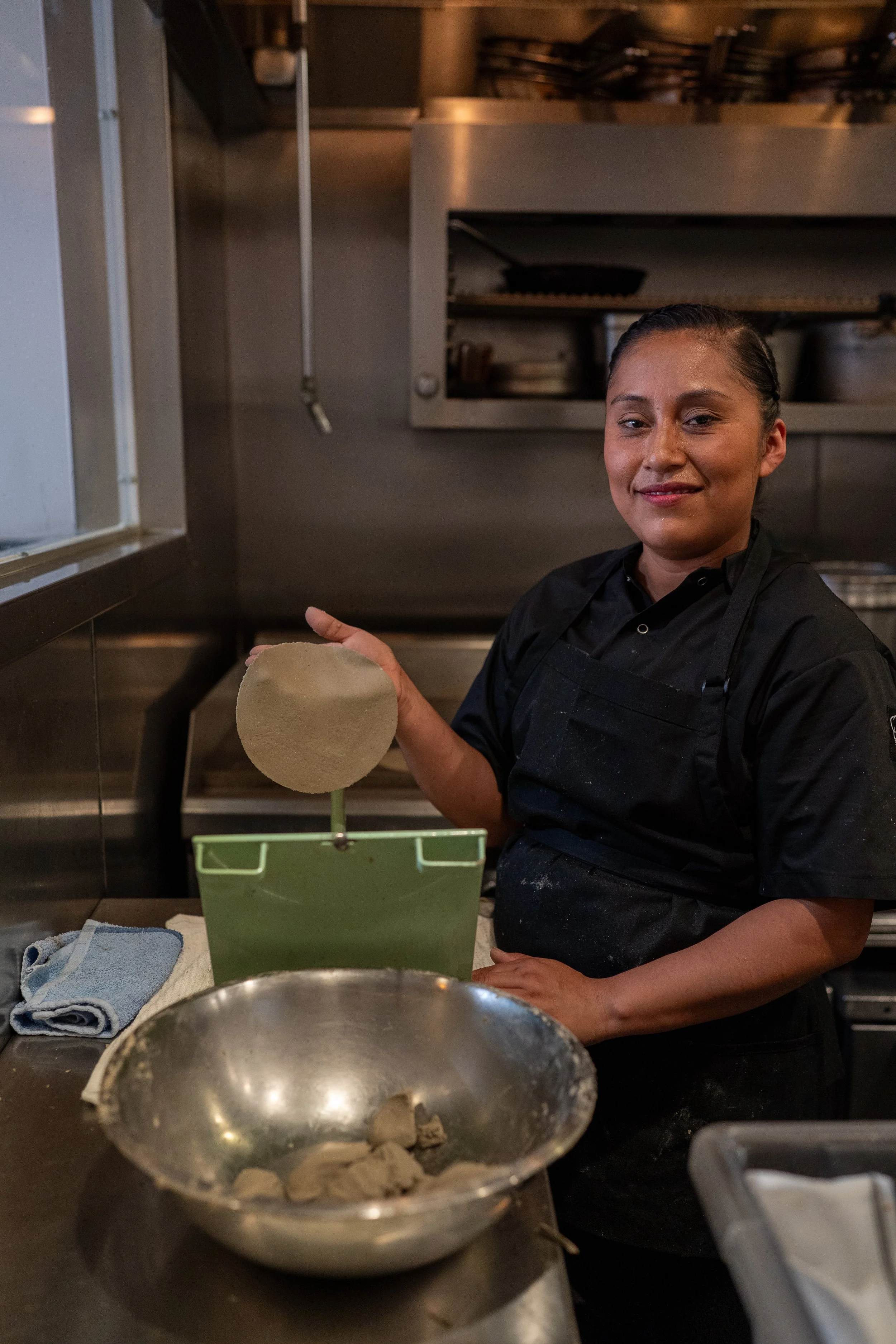 A woman in black chef attire making tortillas by hand using a tortilla press and fresh masa.