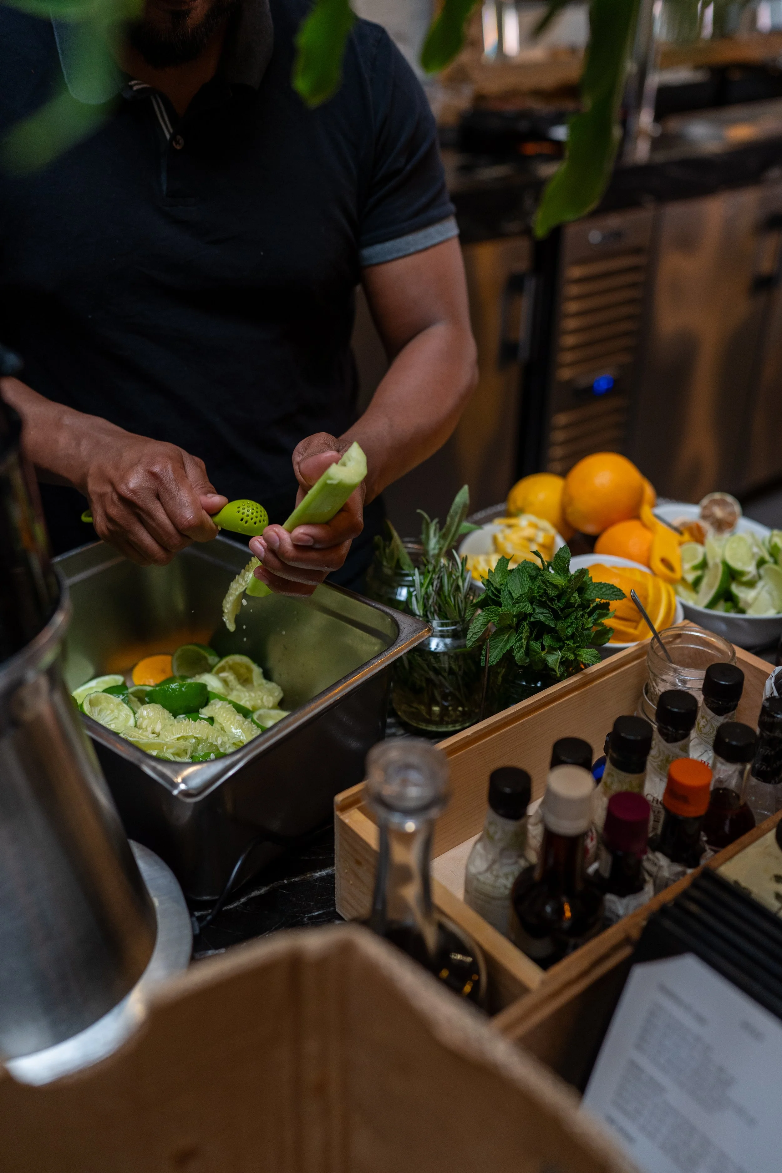 Person slicing lime at a bar or kitchen counter with various fresh fruits, herbs, and spice bottles surrounding him.