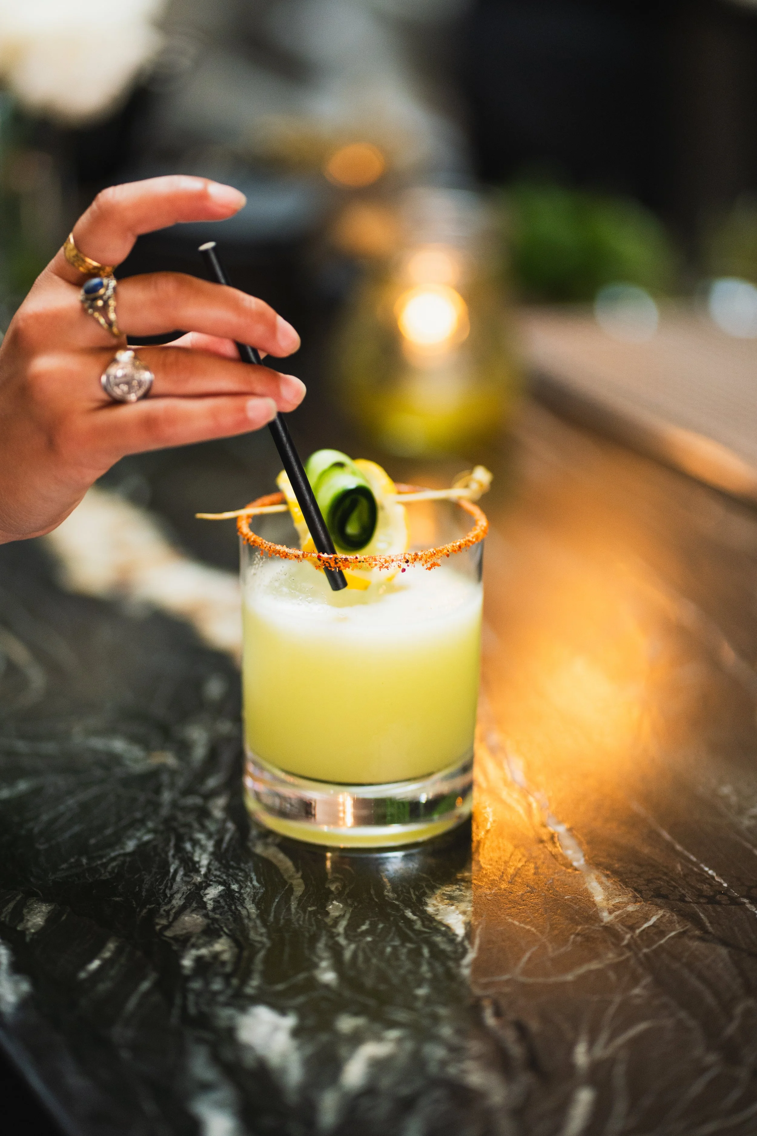 A hand with rings stirring a yellow cocktail with a black straw, garnished with cucumber and chili salt rim, on a dark marble bar.
