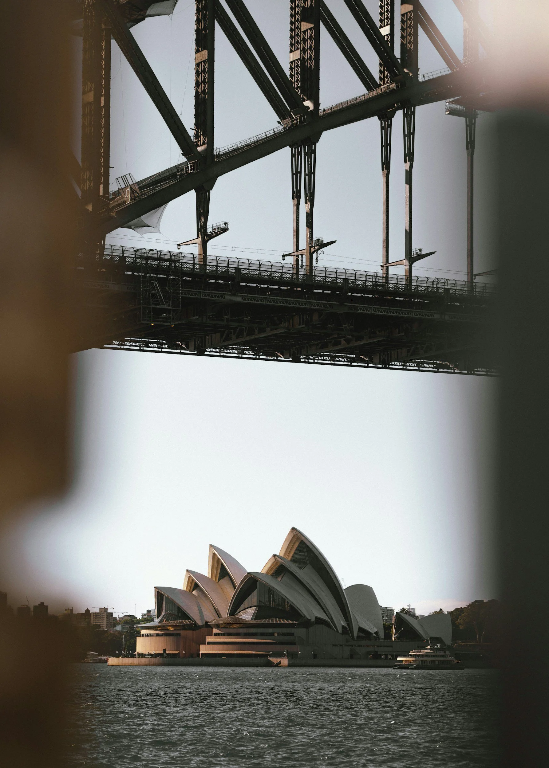 View of the Sydney Opera House framed between parts of a bridge structure, with water in the foreground and city buildings in the background.