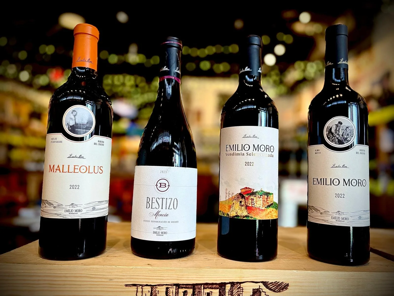 Four bottles of wine on a wooden surface, with blurred colorful background, featuring labels from Emilio Moro winery.