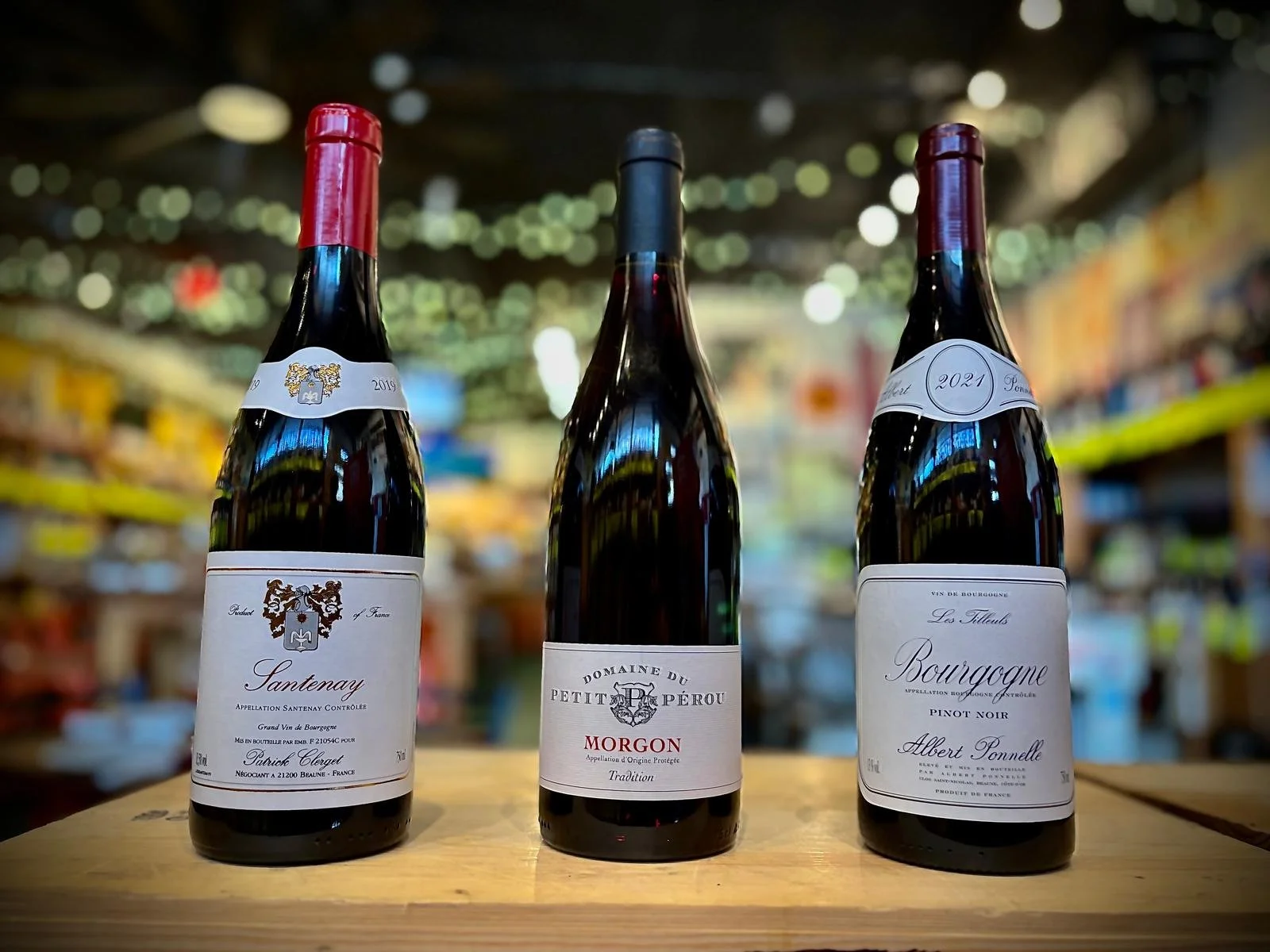 Three bottles of wine on a wooden table in a store, with blurry shelves in the background.
