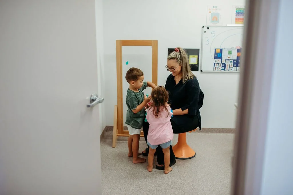 A woman sitting on an orange stool in a classroom with two young children, a boy and a girl, engaging with her near an empty wooden easel.